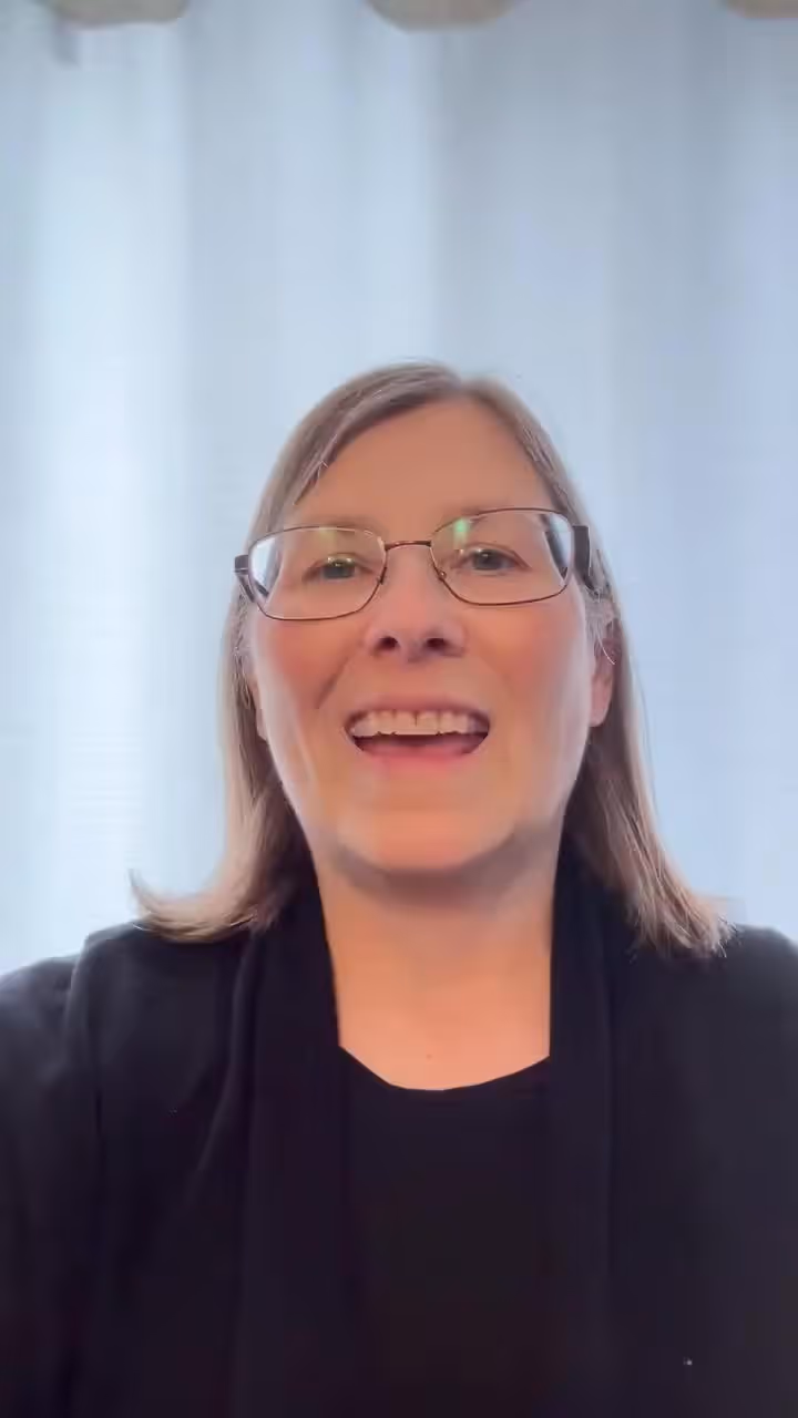 Woman with glasses and black outfit smiling at the camera in a church or hospital setting.