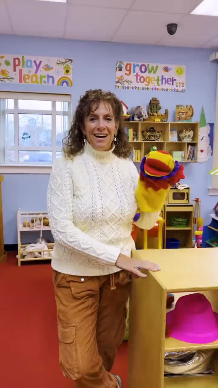 Woman in white sweater holding puppet in educational room with play learn grow together posters.