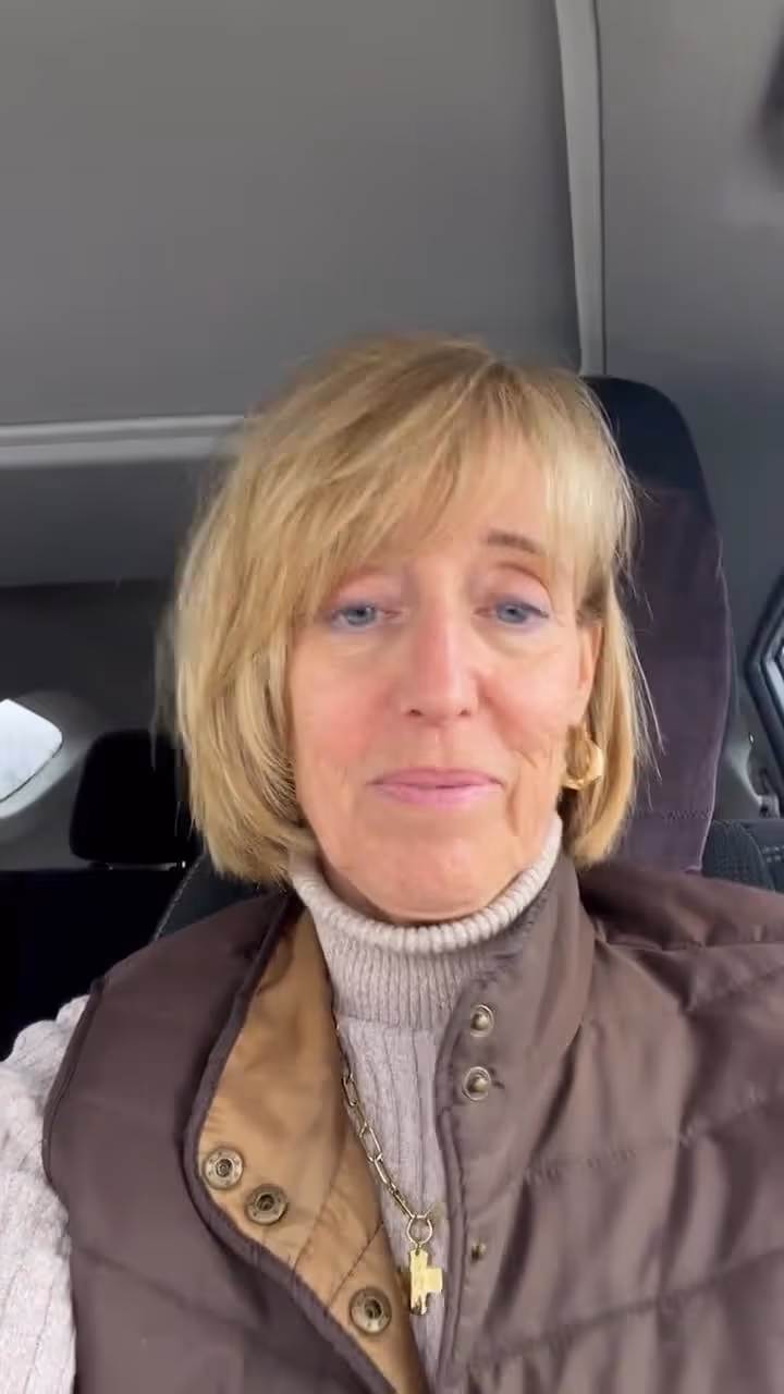 Mature woman with blonde hair sitting in a vehicle wearing a brown vest and turtleneck.