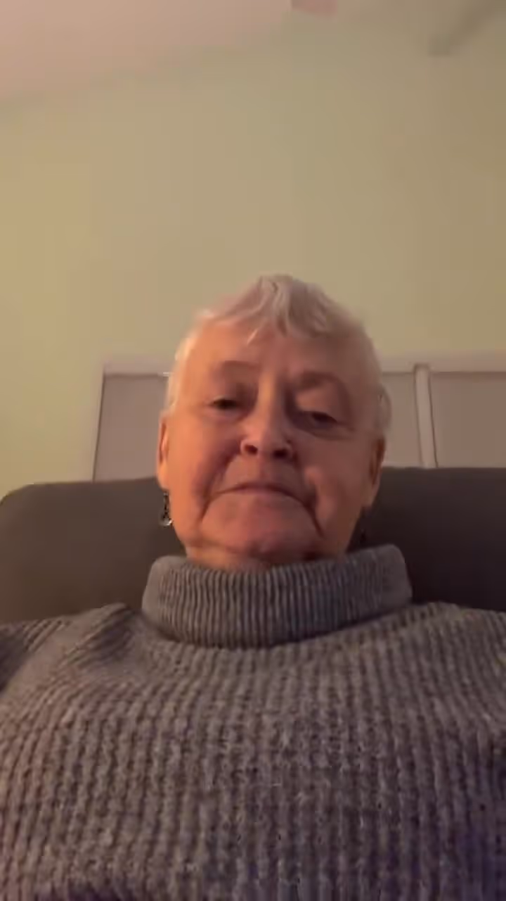 Elderly woman with short white hair sitting on a couch wearing a gray turtleneck sweater.