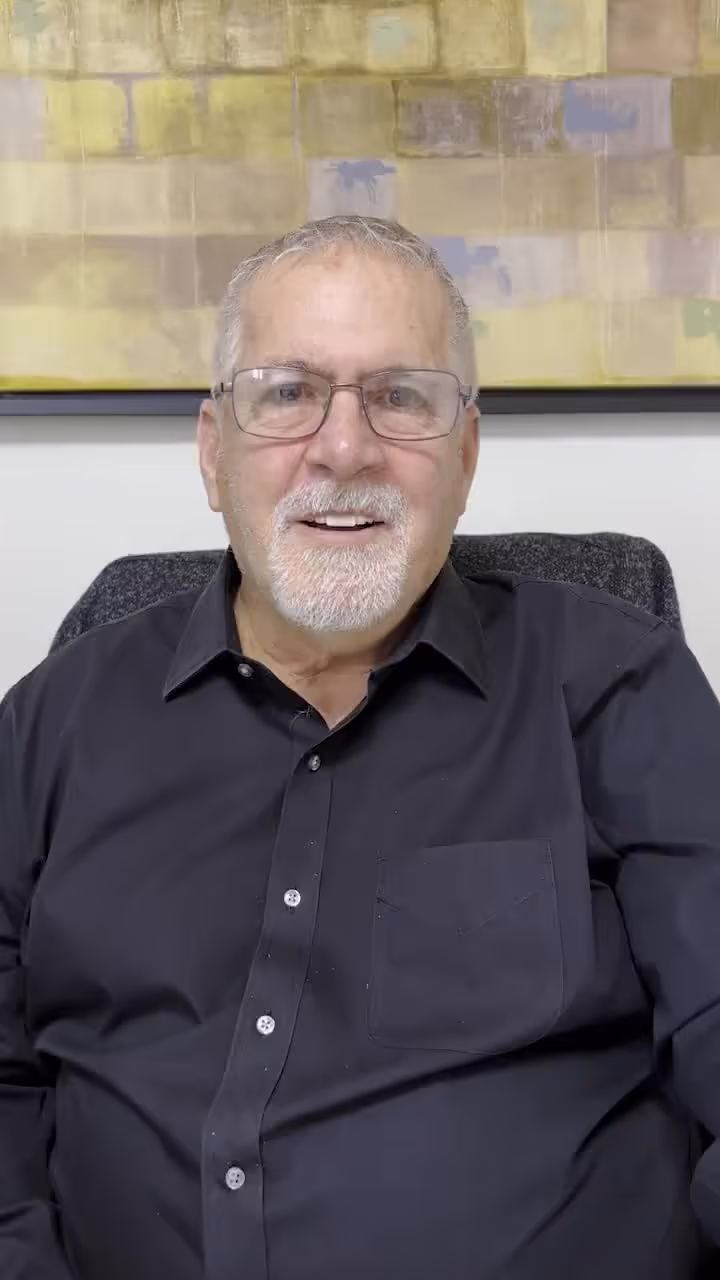 Older man with glasses and goatee sitting in office, possibly hospital or church setting.