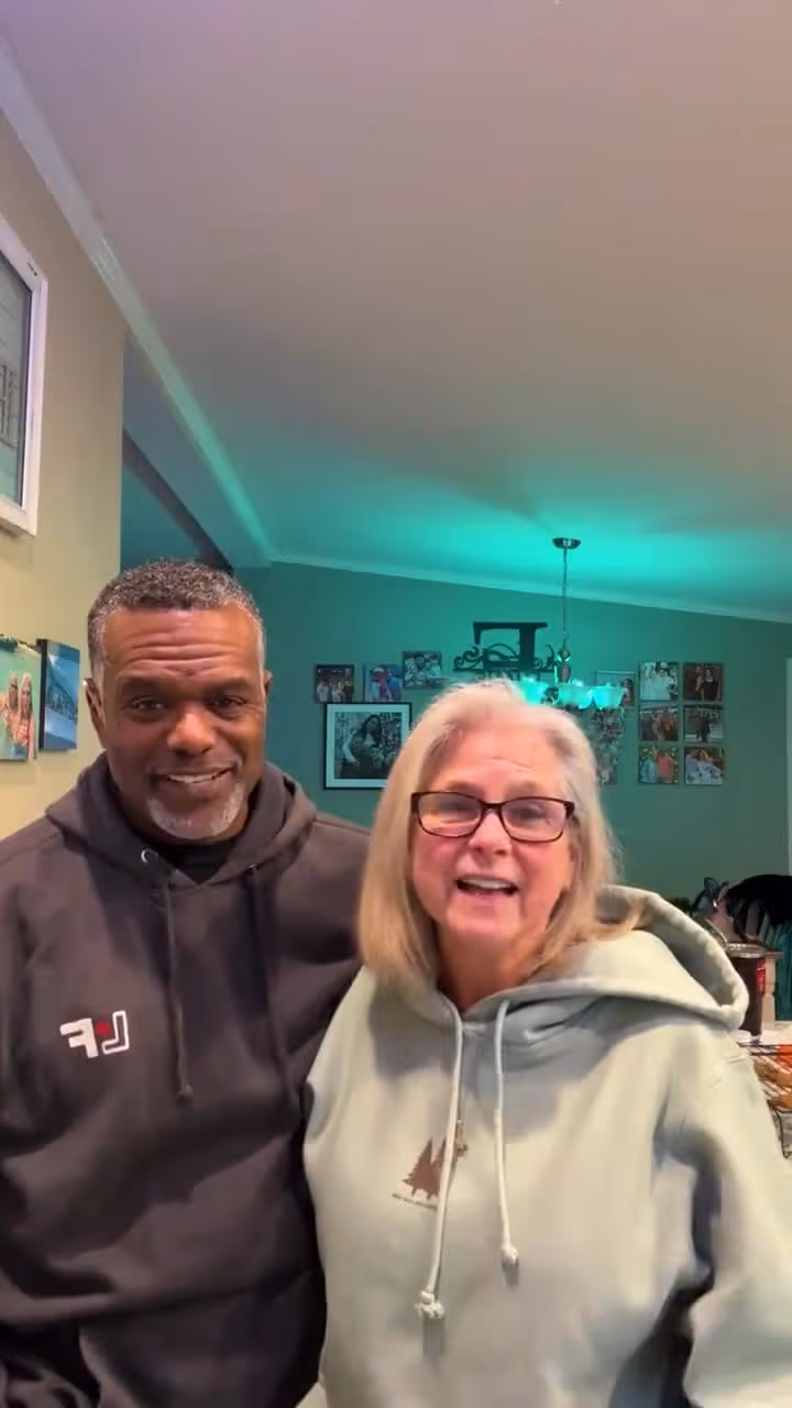 Smiling middle-aged couple standing together in a cozy indoor setting with family photos behind them.