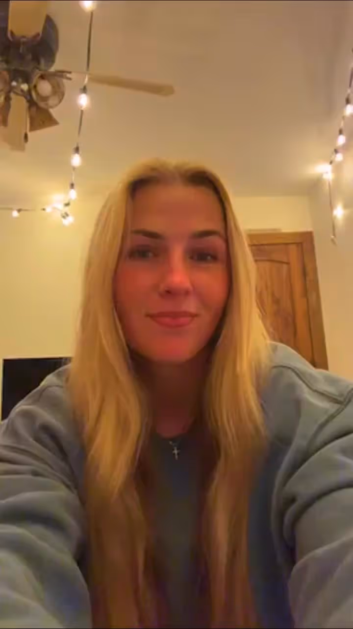 Blonde woman wearing cross necklace smiling at camera indoors with string lights and fan visible.