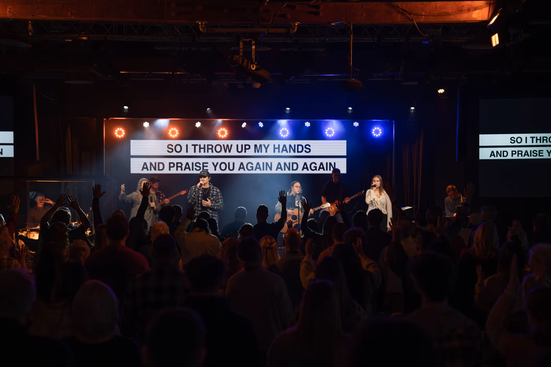 Worship team performs on stage with raised hands and lyrics displayed behind them in church setting.