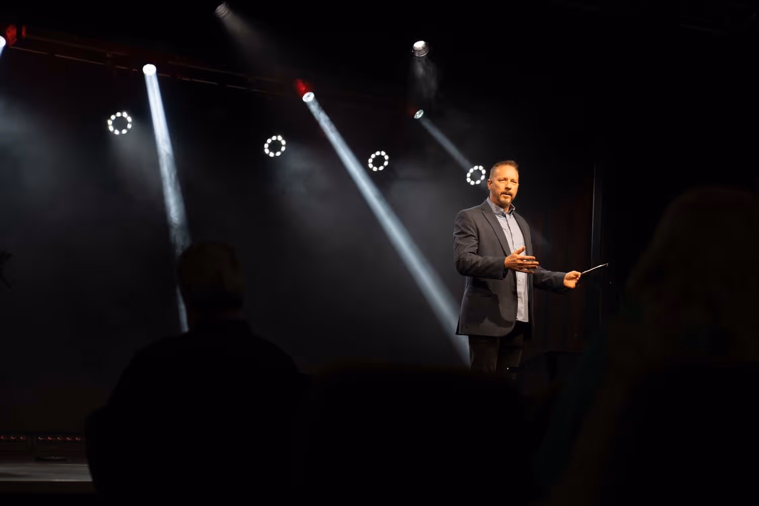 Man in suit speaking on stage with spotlights shining down on him from above.