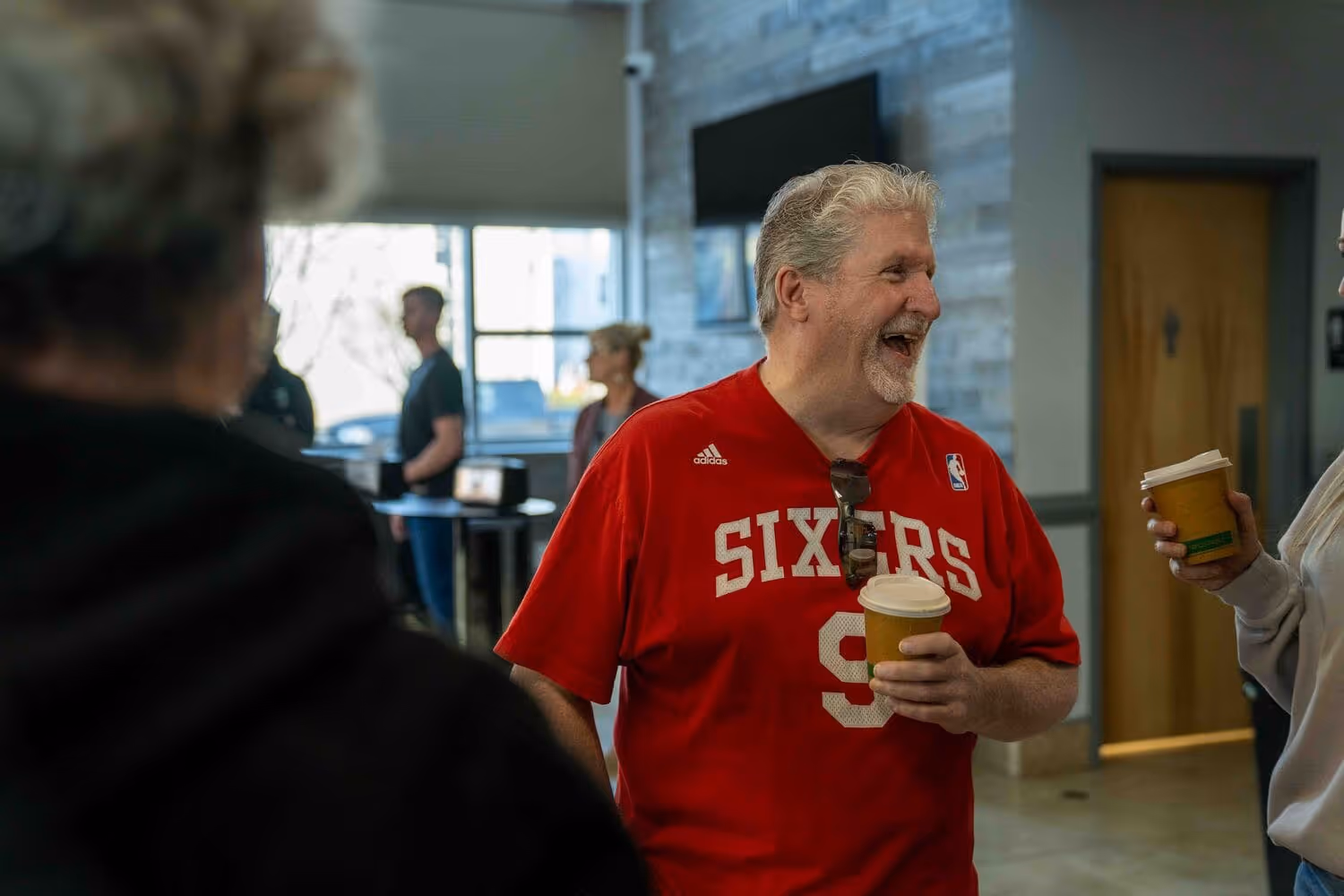 Man in red Sixers jersey holding coffee cup talking to people in church or hospital setting.