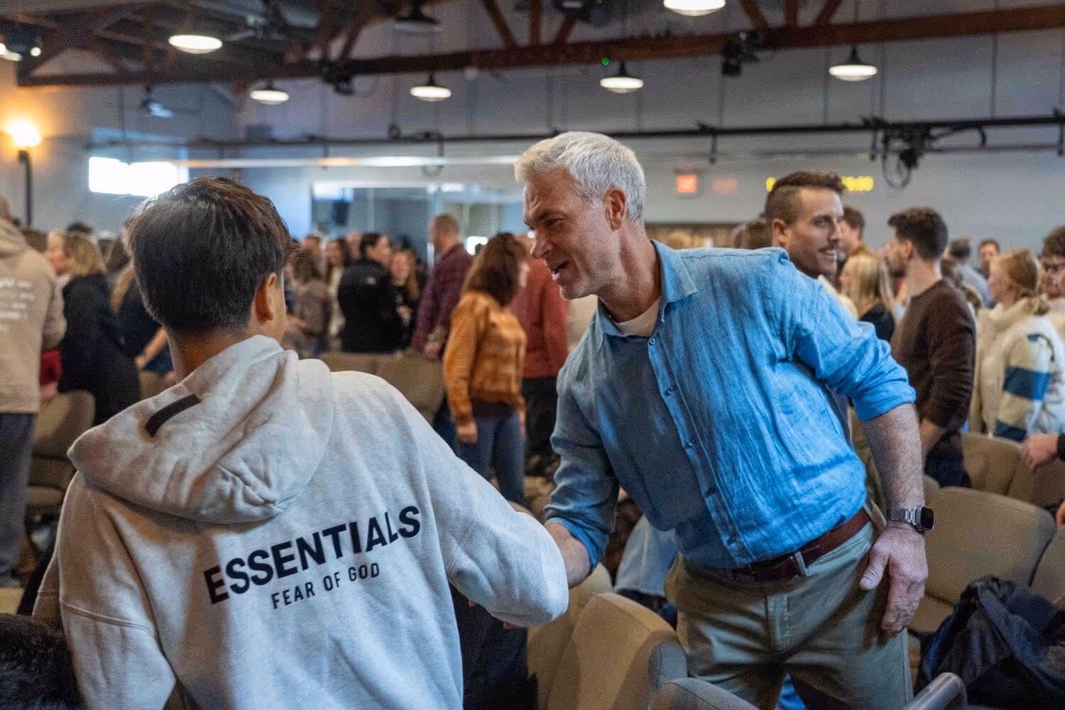 Man and youth interacting at church event with crowd in background, wearing religious apparel.