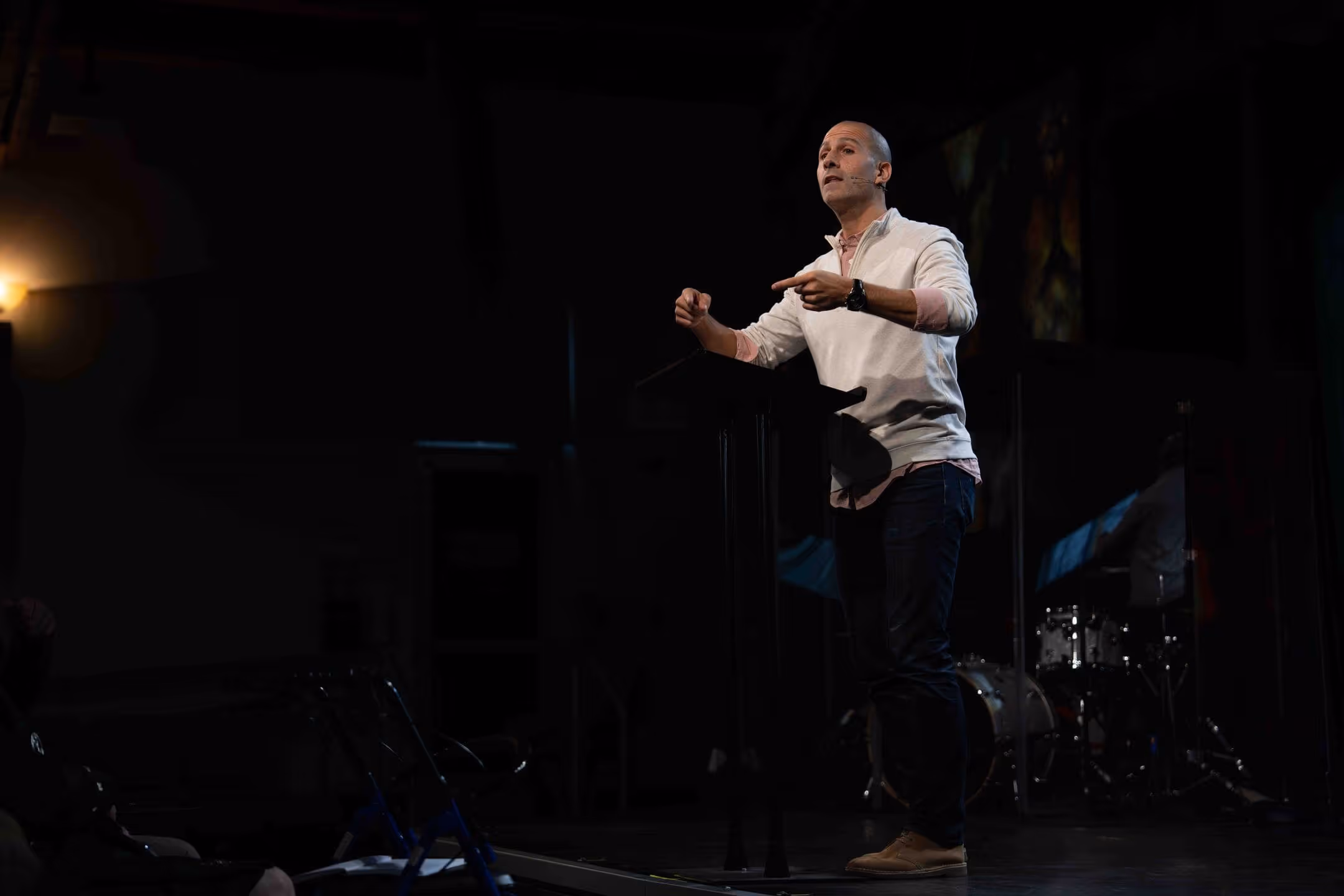 Man speaking at a church event with a microphone and drum set in the background.