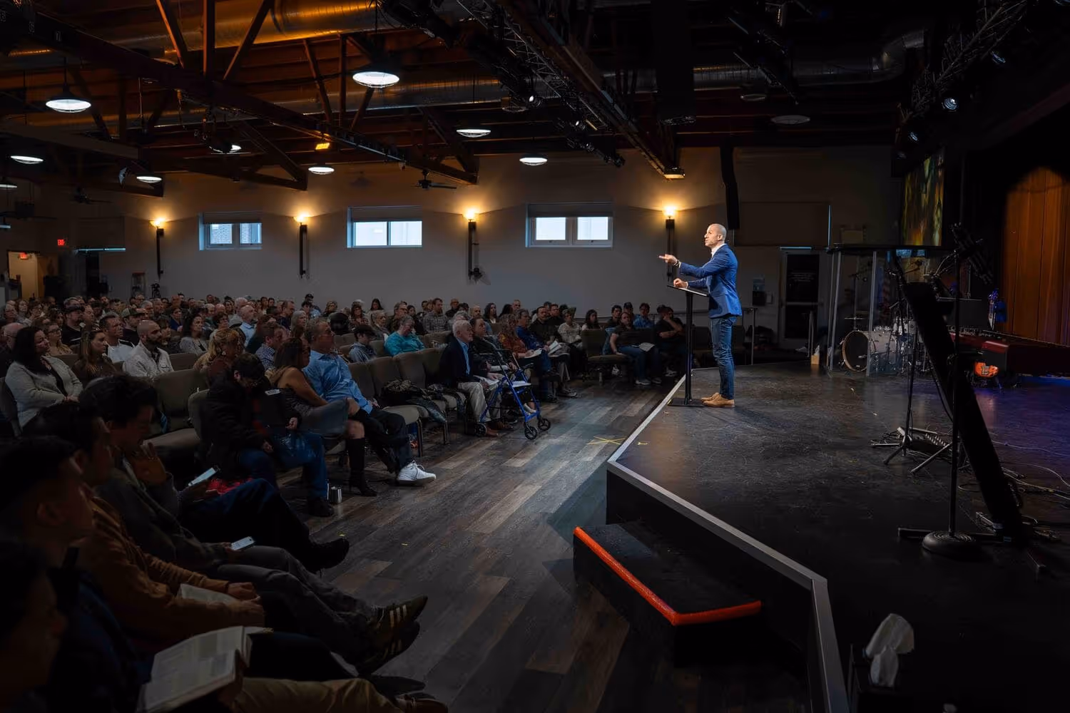 Man in blue suit speaks to a large crowd in a church auditorium setting.