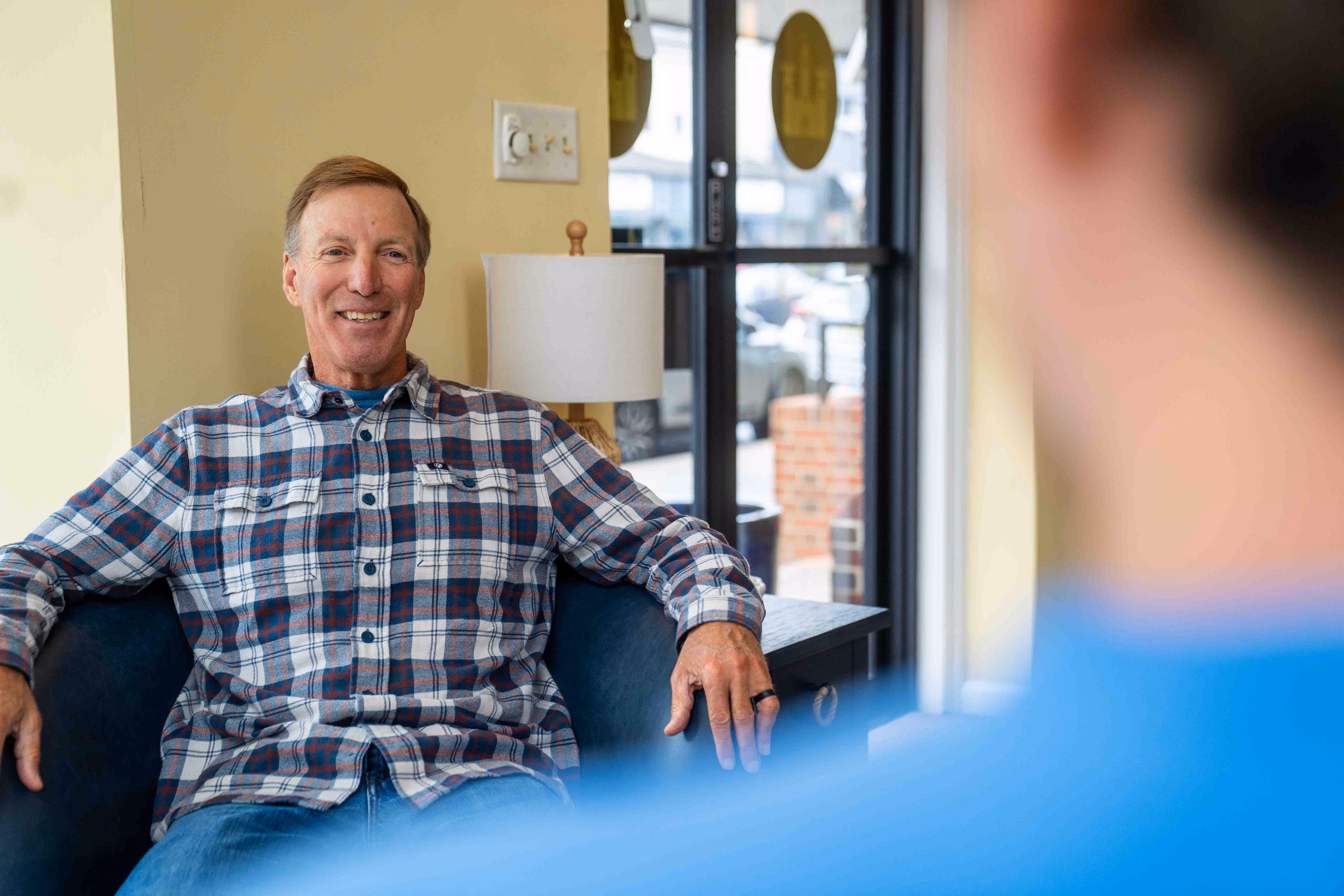 man sitting in chair at hospital or church talking to someone in blue shirt.