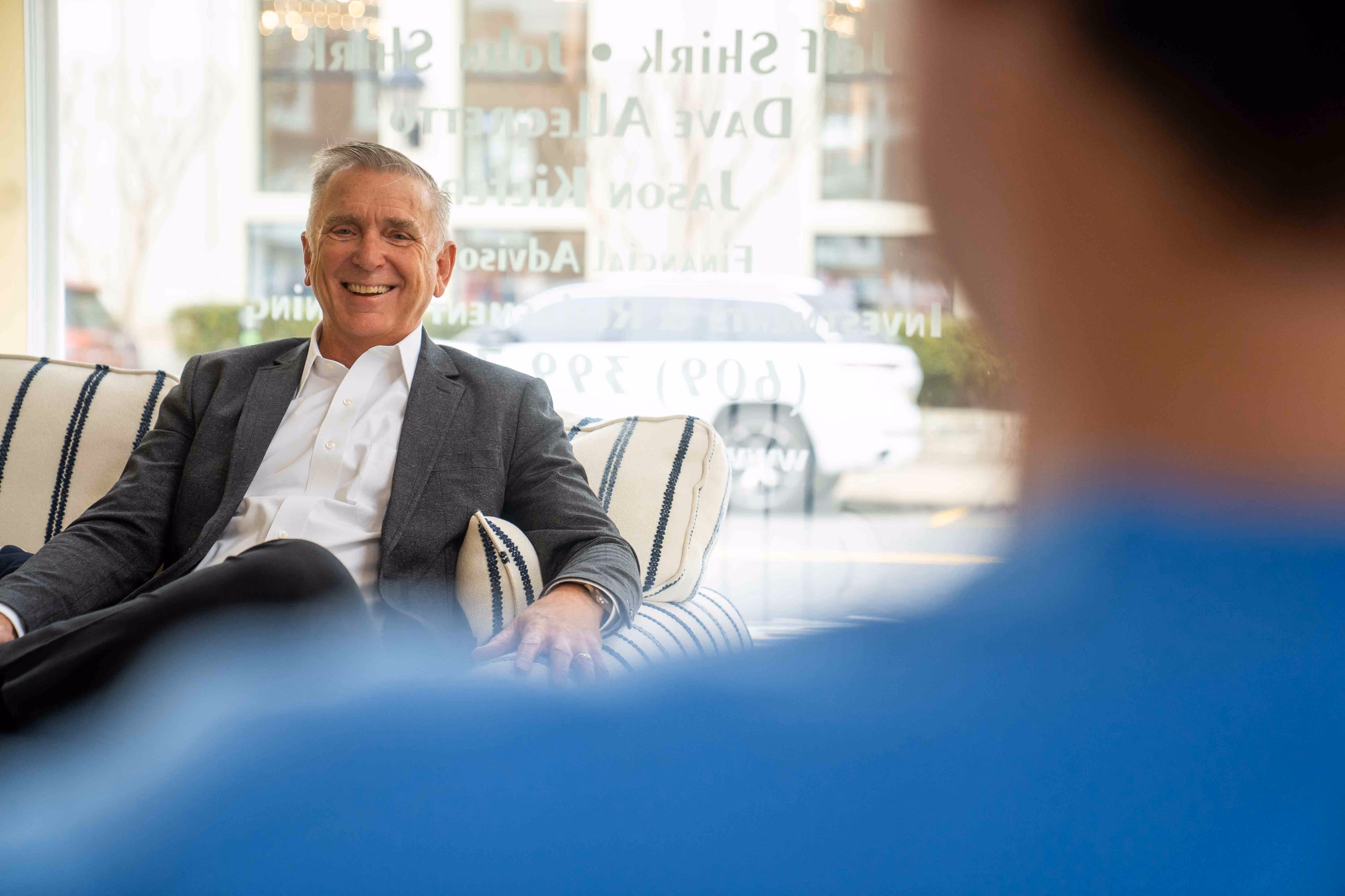 Smiling man in suit sitting on couch talking to person in blue shirt at church or hospital.