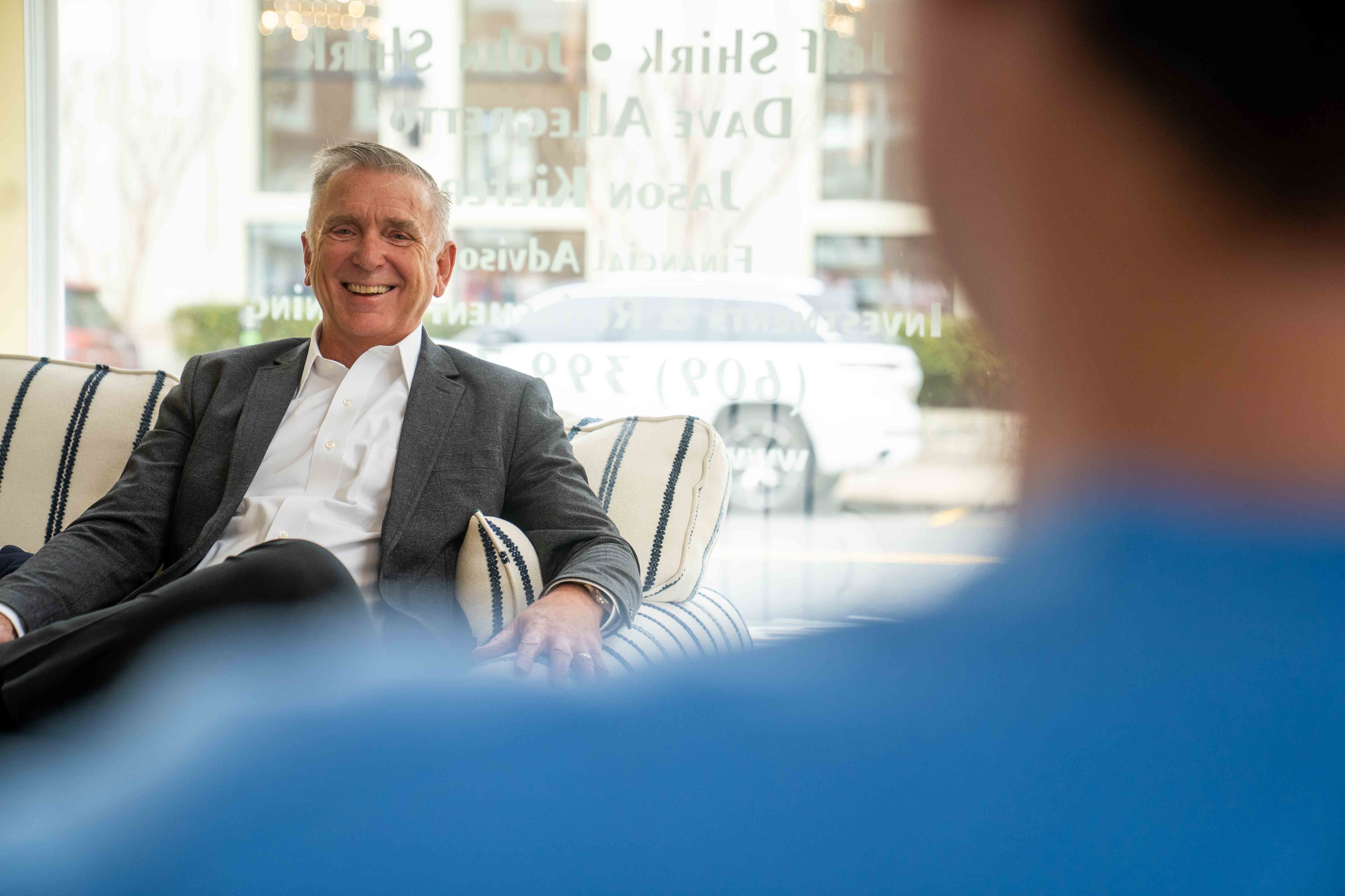 Smiling man in suit sitting on couch talking to person in blue shirt at church or hospital.
