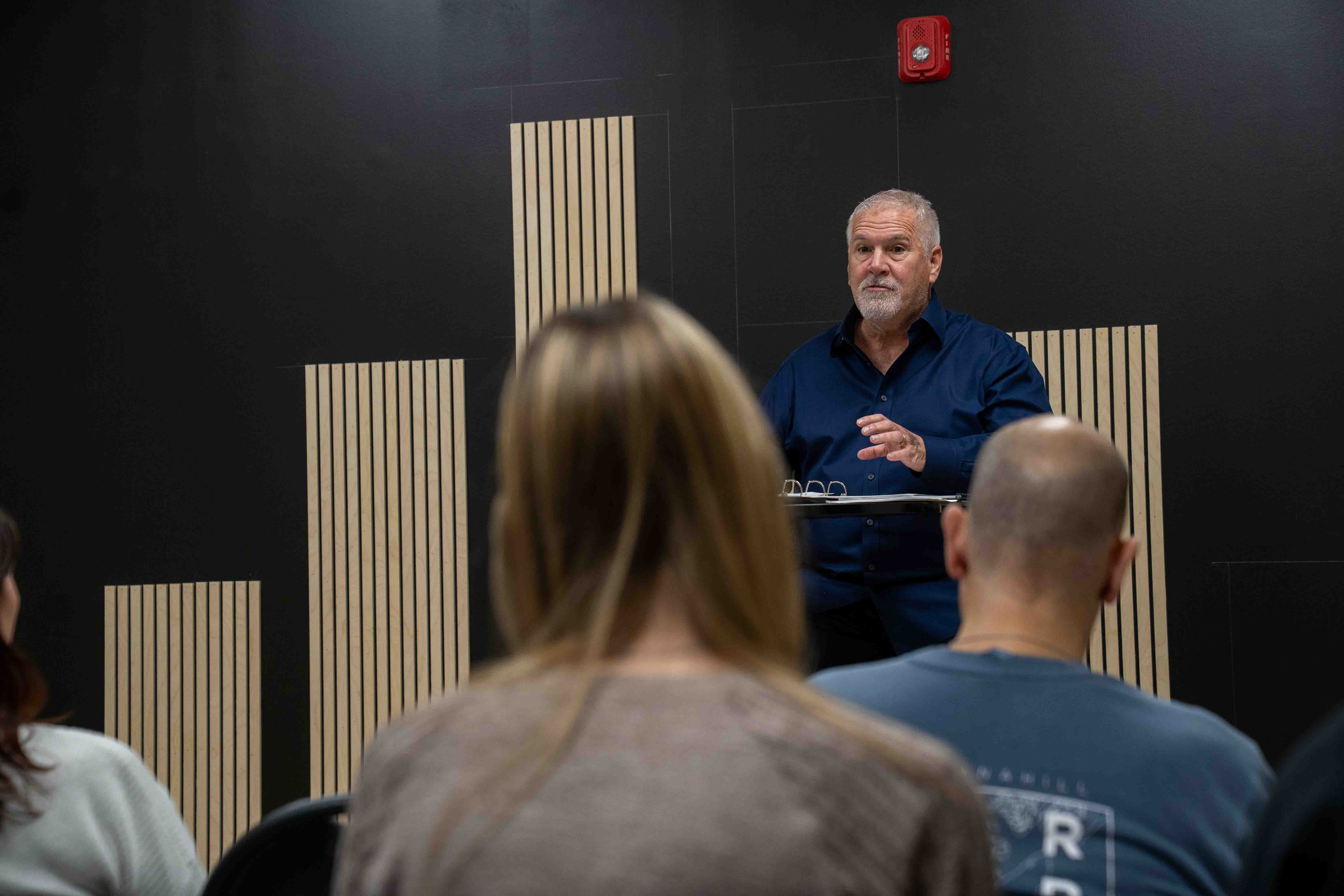Man speaking at church or hospital event, addressing audience from behind a lectern or table.
