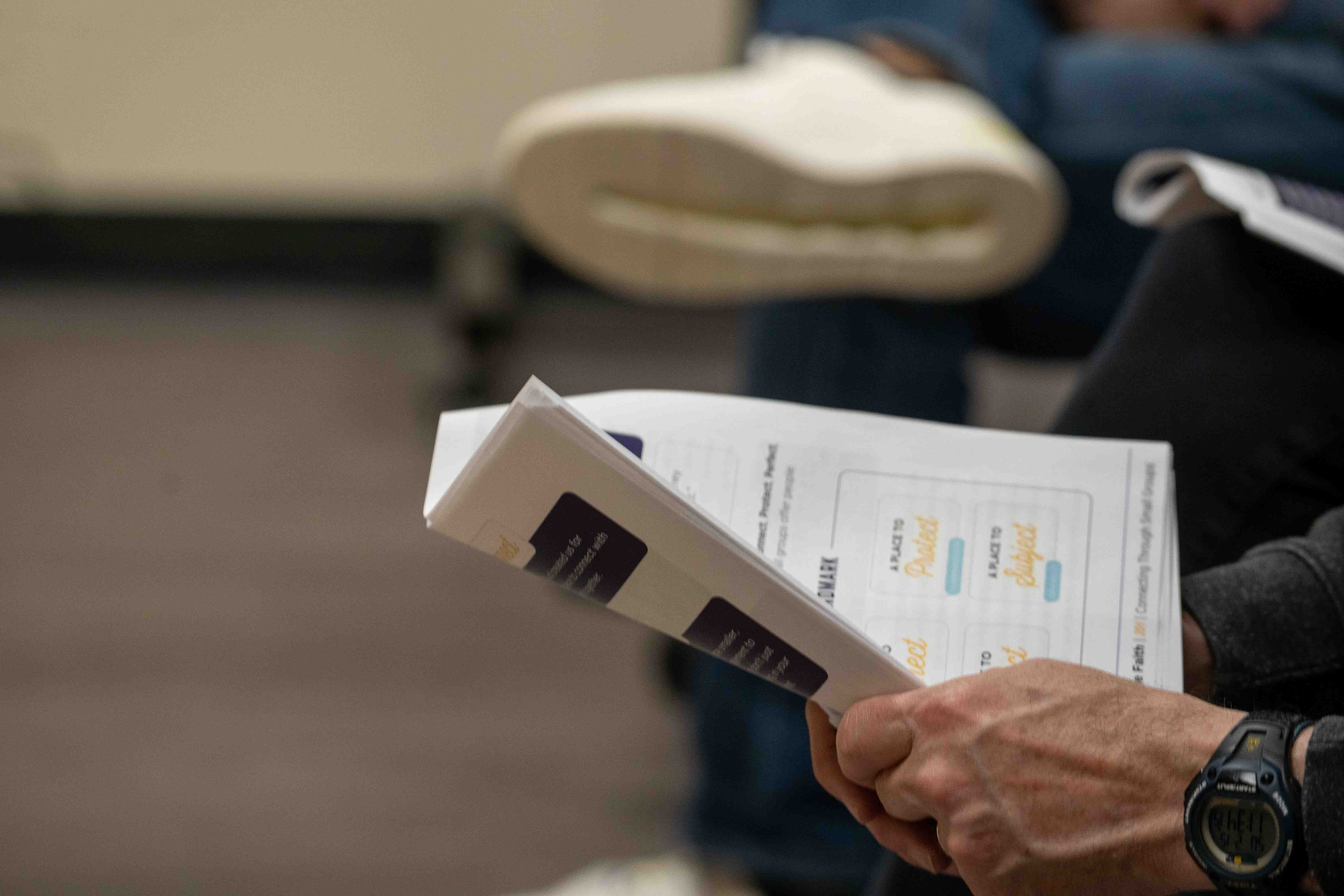 Person holding church program or hospital brochure with watch on wrist in meeting or service setting.