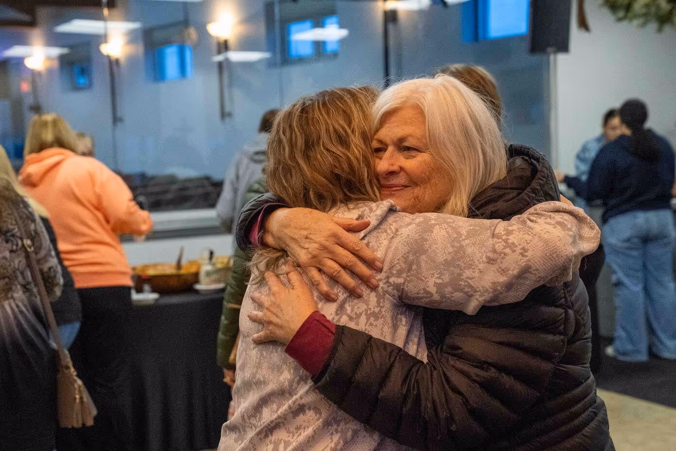 Two women embracing warmly in a church setting surrounded by people.