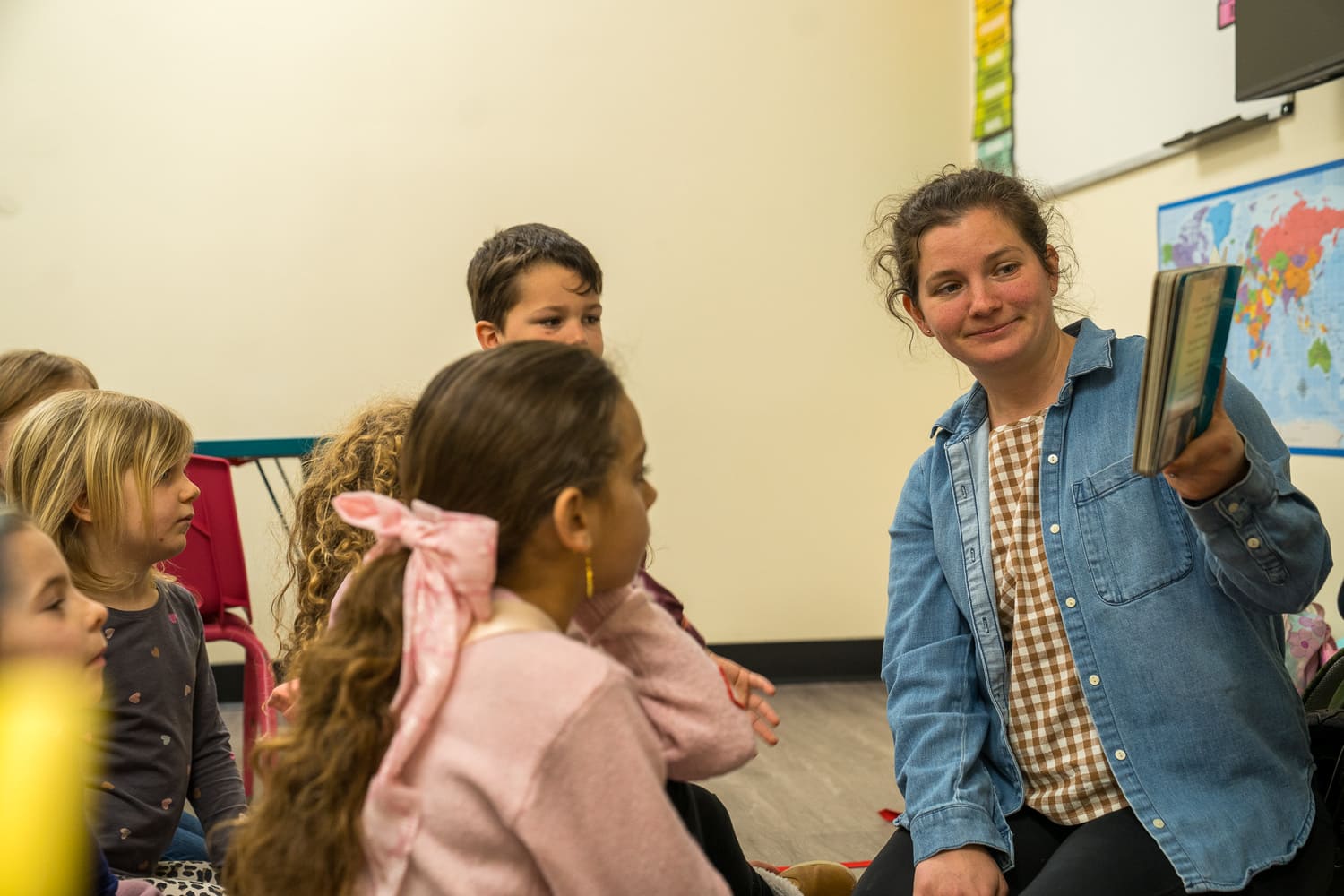 Woman reads to children in classroom, likely hospital or church program, with world map background.