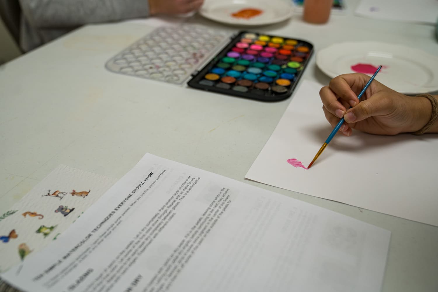 Watercolor painting class with hand holding brush, paints, and techniques guide on table at church or hospital.