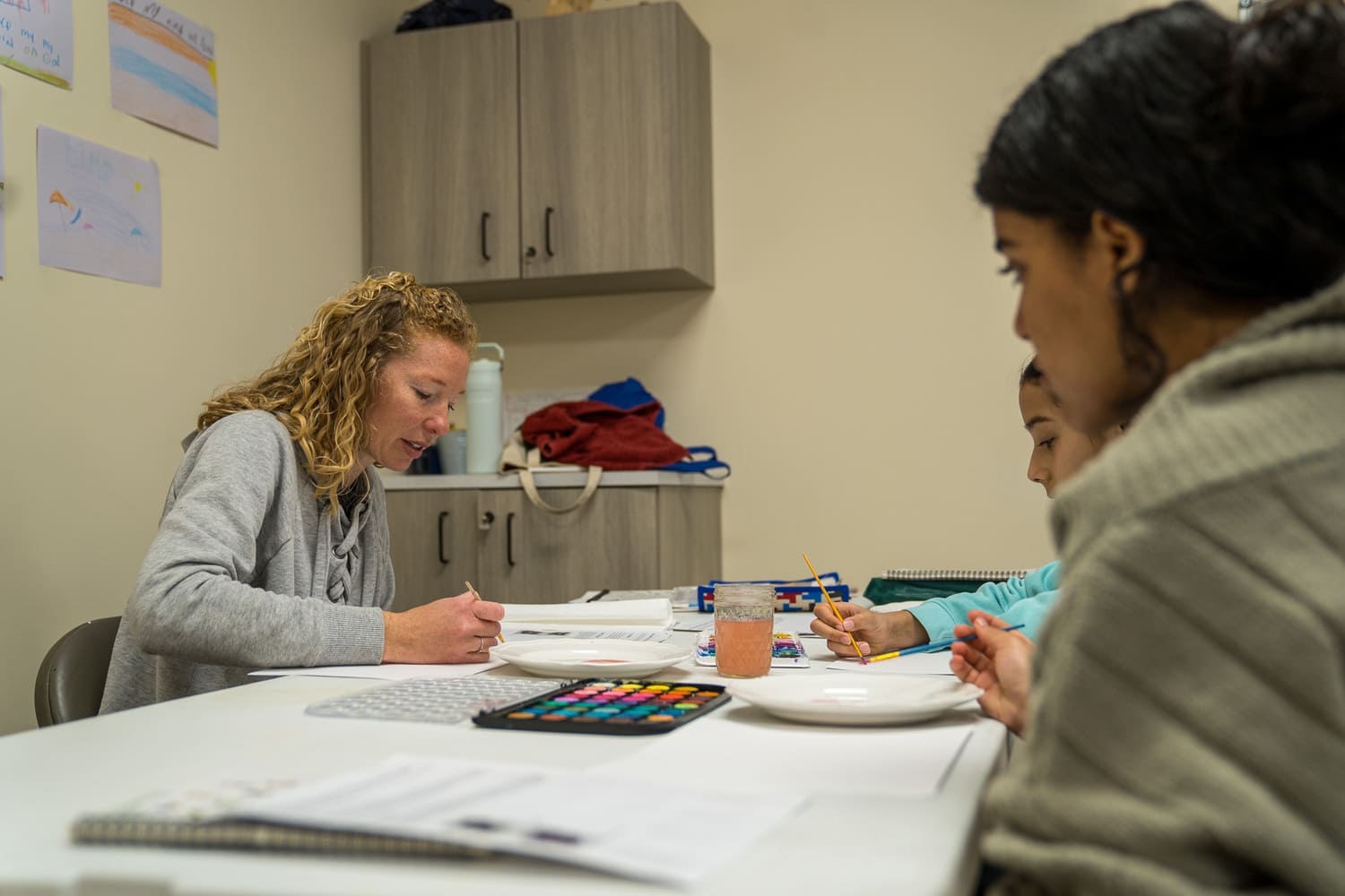 Woman and children engaged in creative activity with paint and paper at a table indoors.