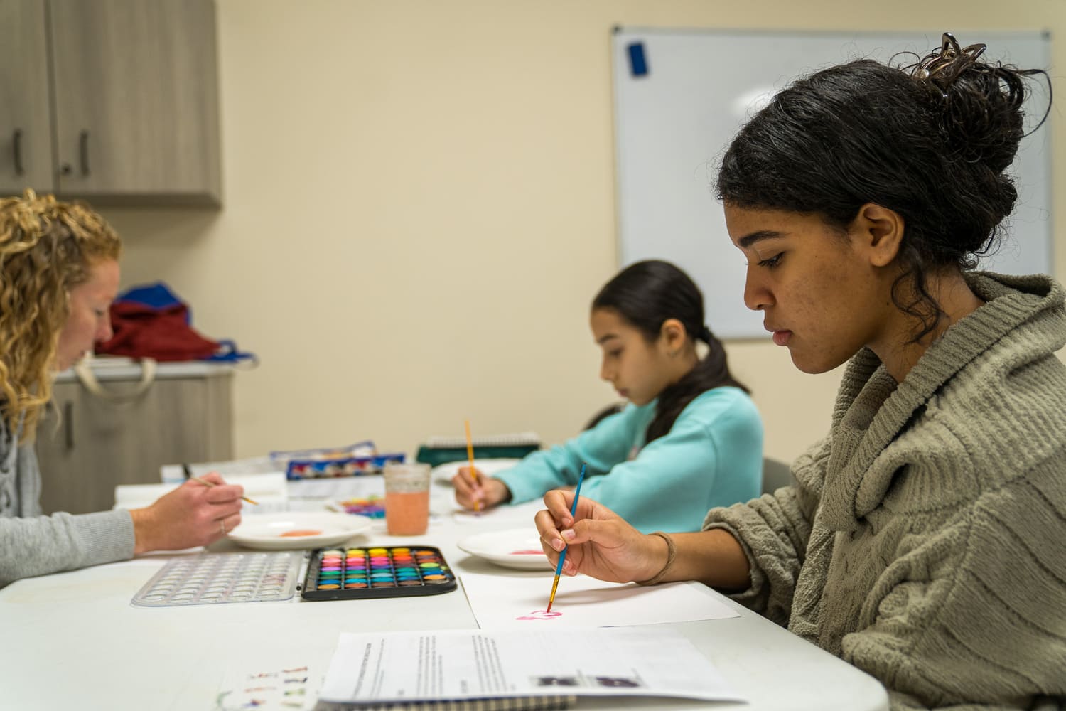 Women painting with watercolors at a table in a classroom or hospital setting near a church.