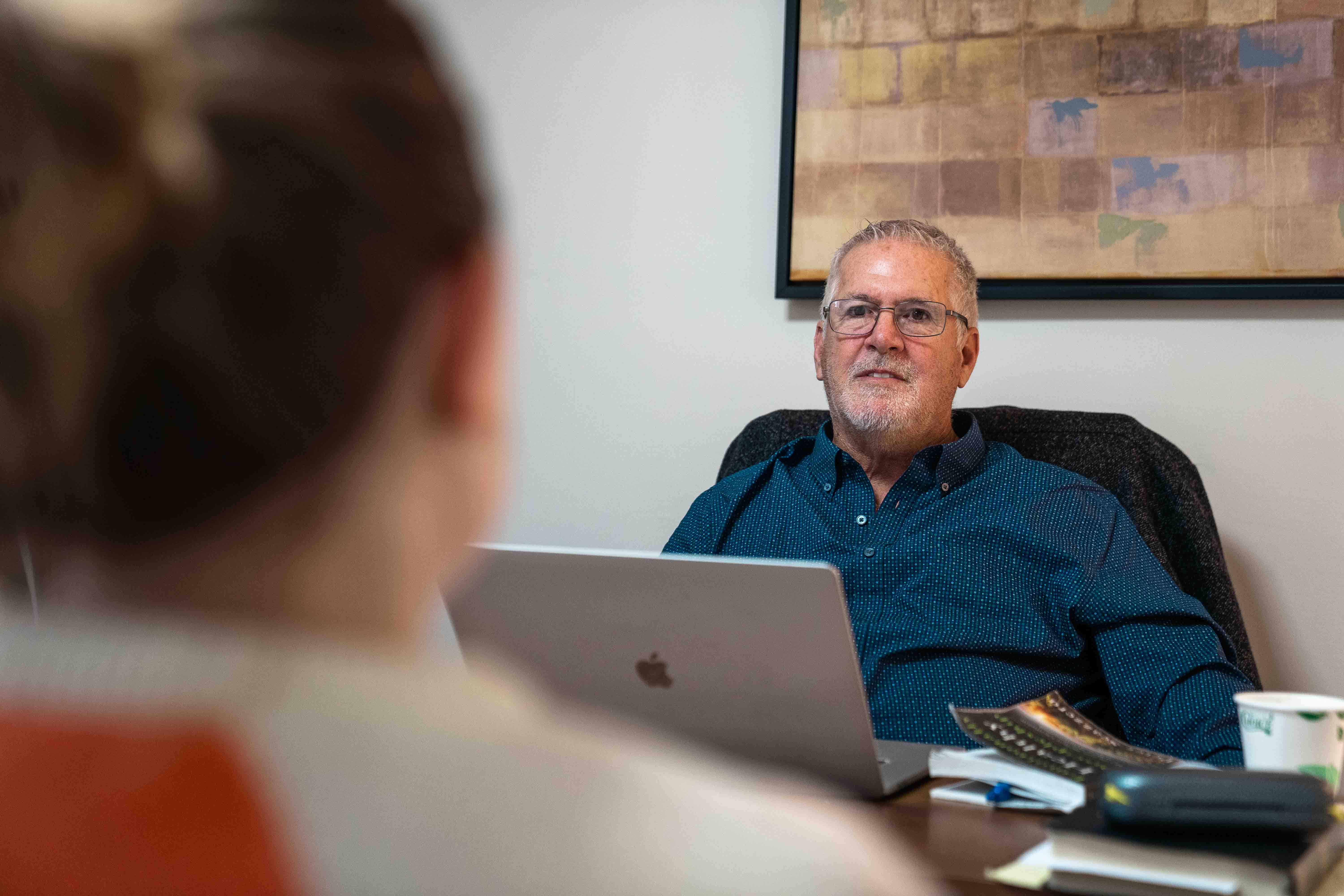 Man in office with laptop, talking to woman across desk in church or hospital setting.
