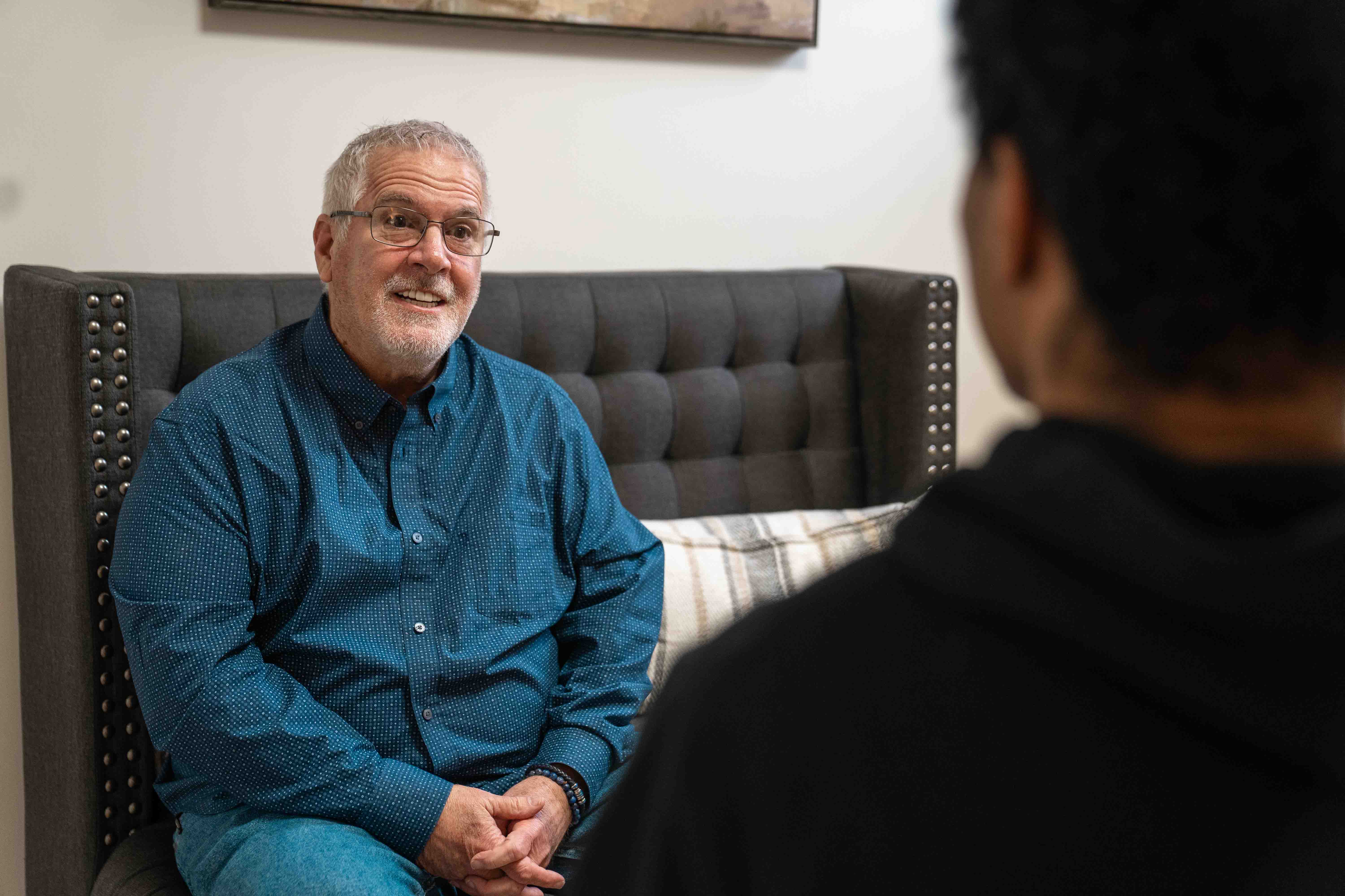 Older man in blue shirt sitting on couch talking to someone in a therapy session.