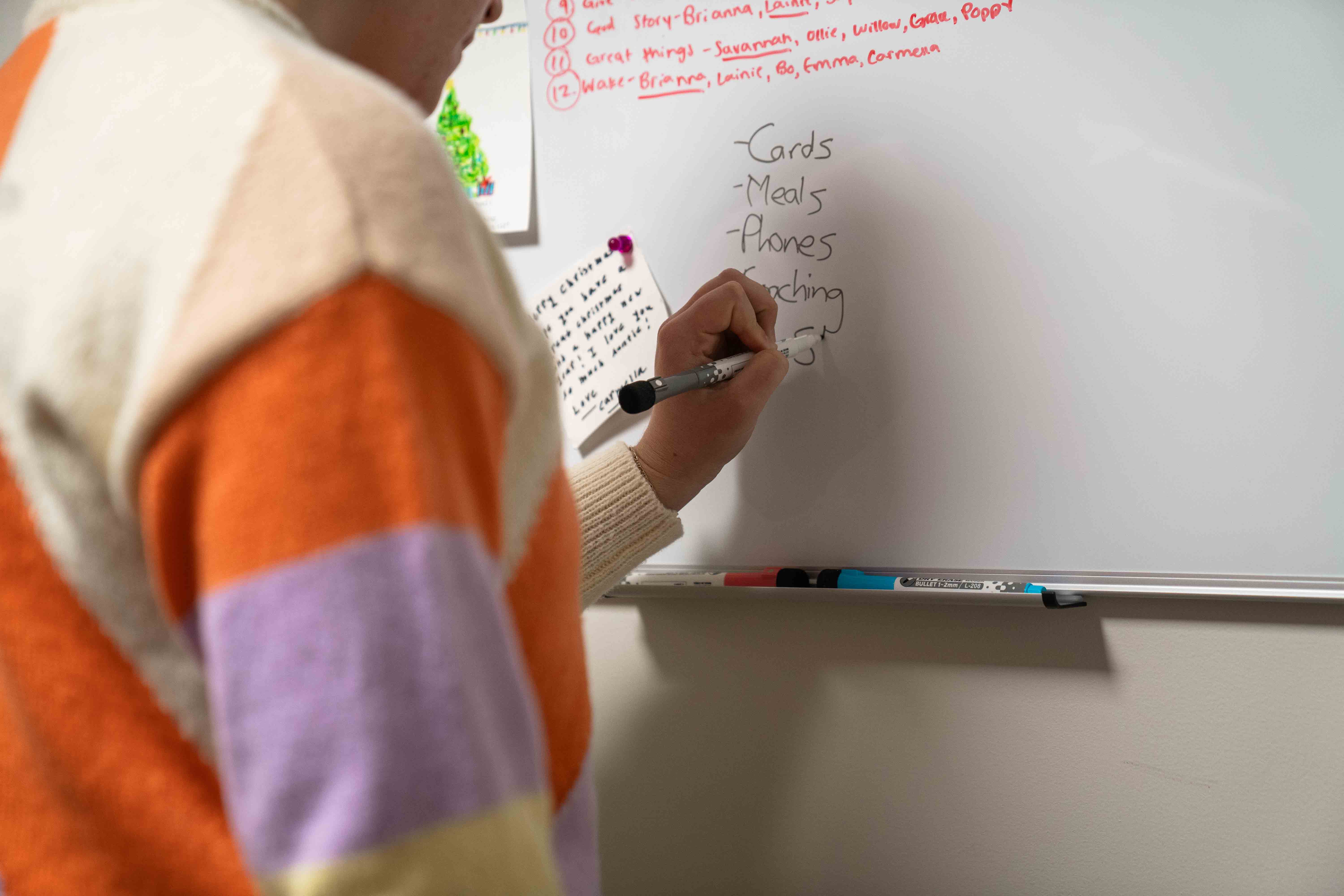 Person in a colorful sweater writing a list on a whiteboard with items including cards, meals, phones, and coaching.
