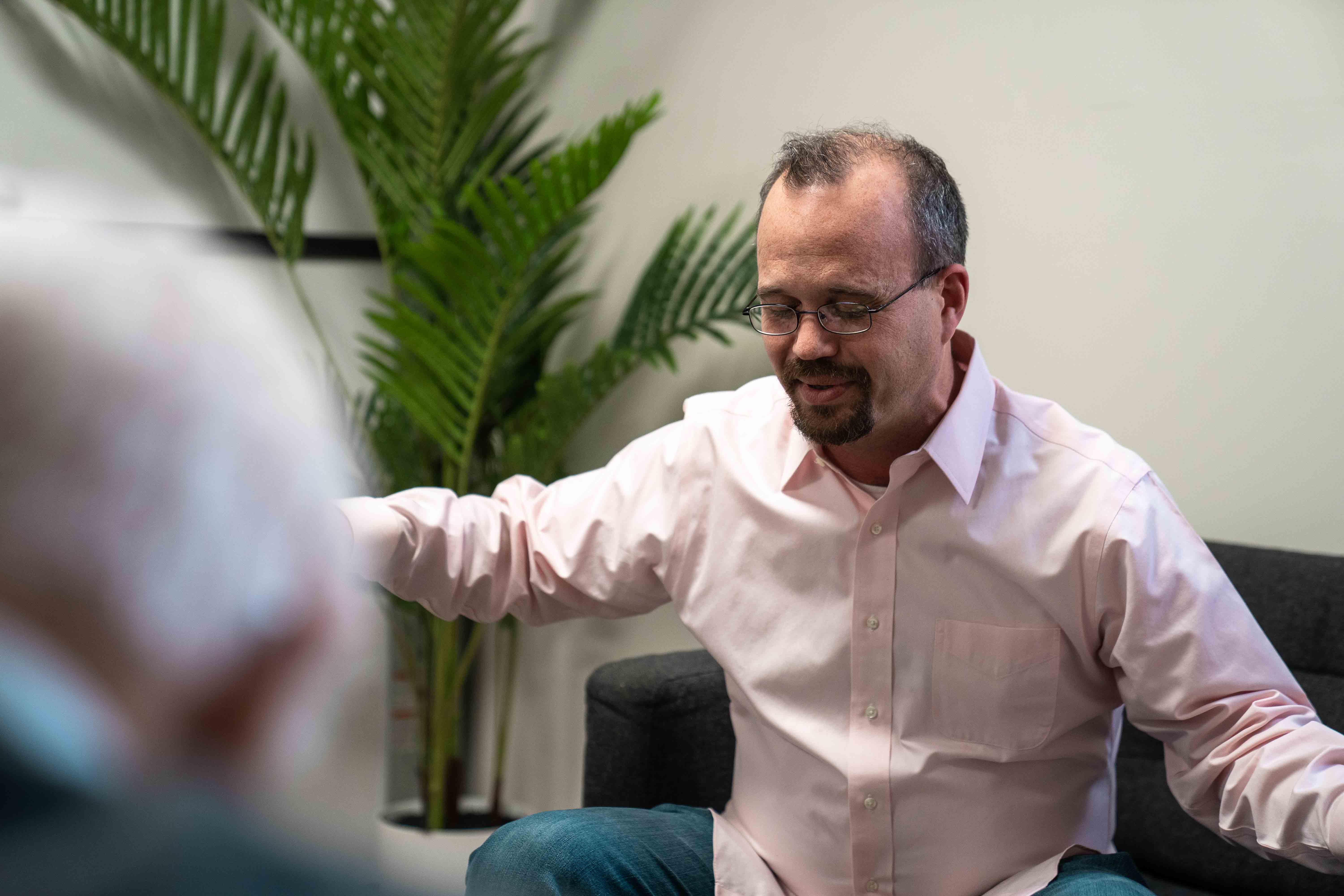 Man in glasses and pink shirt sitting on a couch with arms extended, talking to an older person.