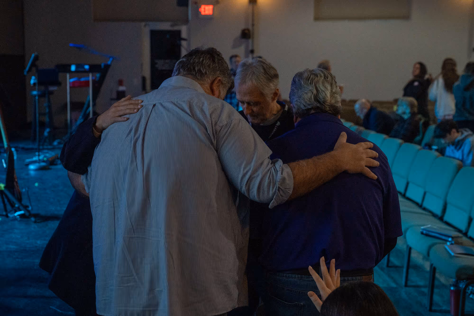 Men embracing in prayer at a church service with rows of chairs and audience.