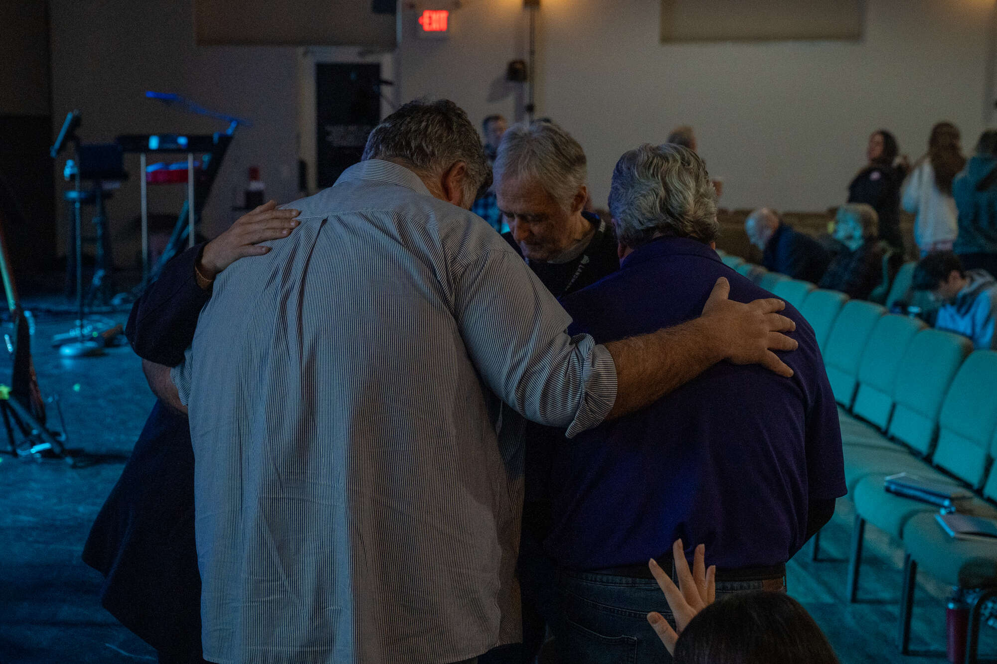 Men embracing in prayer at a church service with rows of chairs and audience.