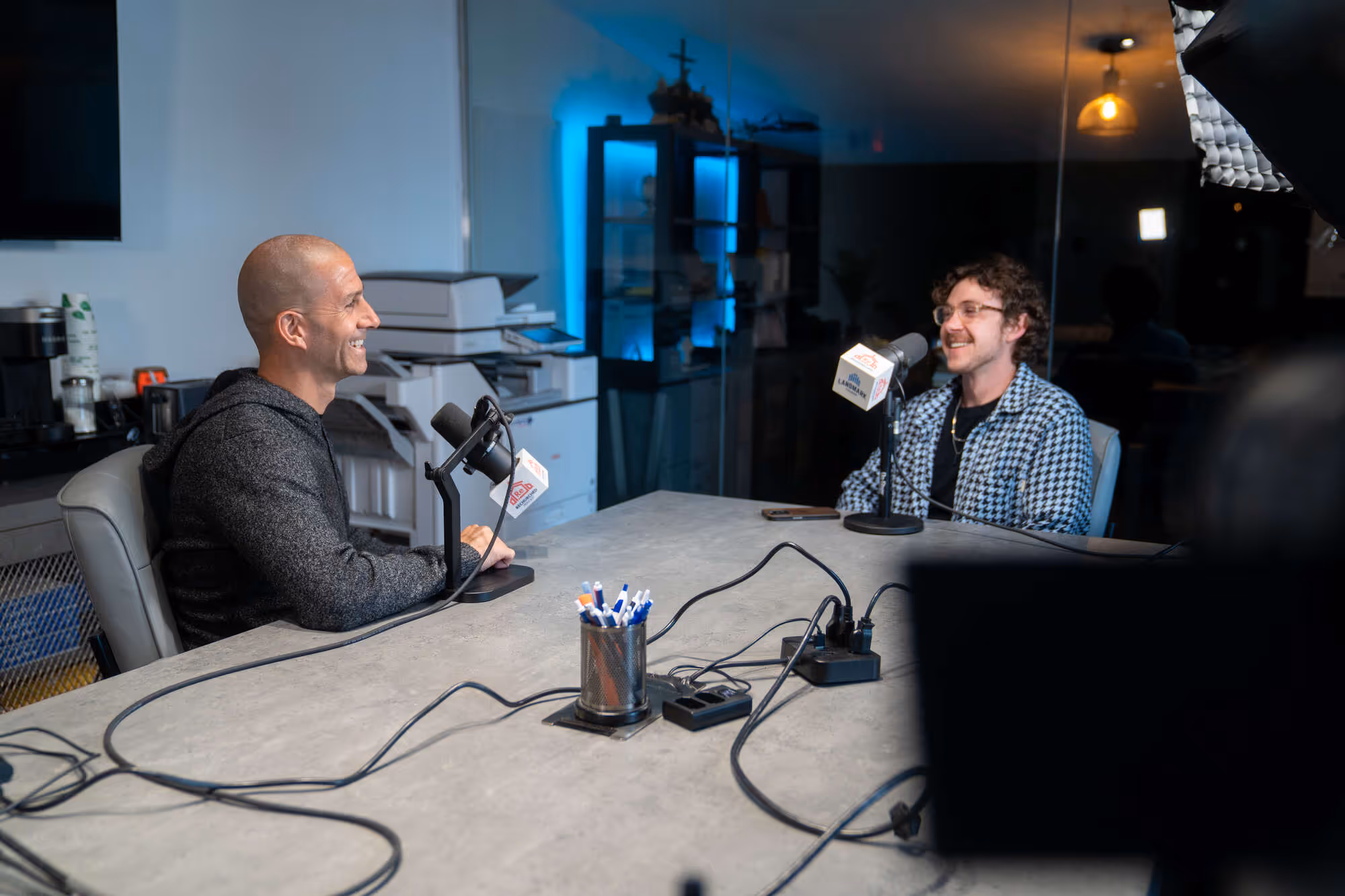 Two men sitting at a table with microphones in a church or hospital media room setting.