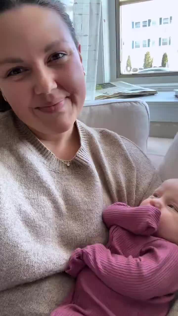 Woman holding newborn baby on couch with hospital and church buildings visible outside window.