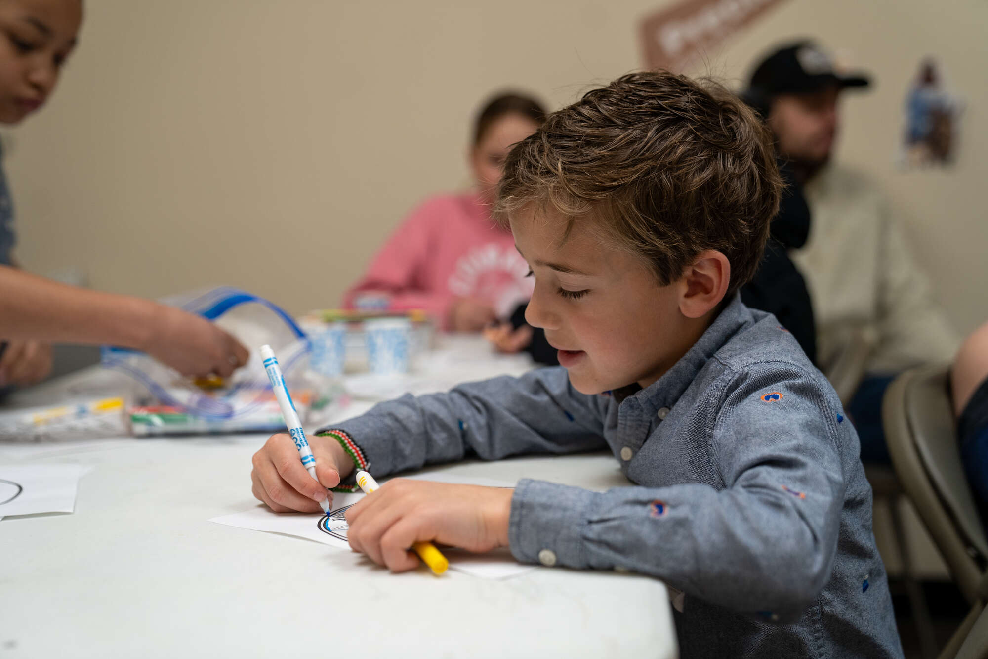 Children coloring at church event with markers and paper on a table surrounded by people.