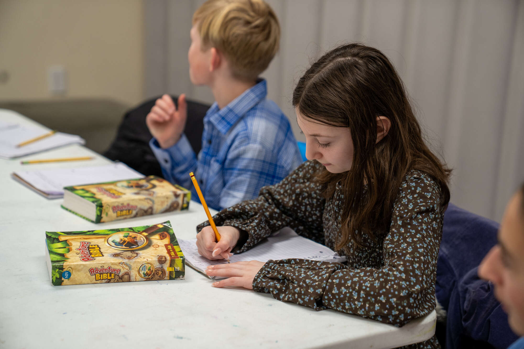 Children writing with bible and notebook on table in church or hospital setting.