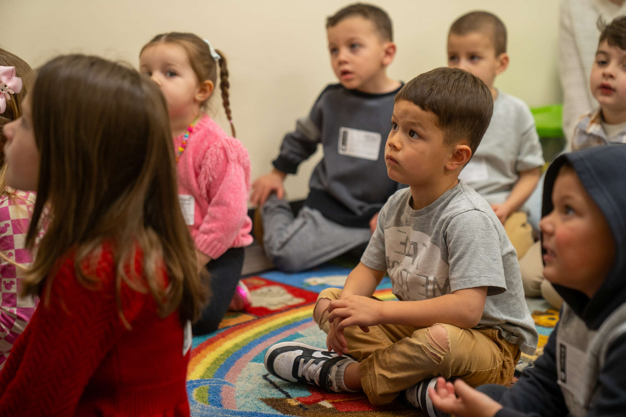 Children sitting on colorful rug in classroom or church program with name tags.