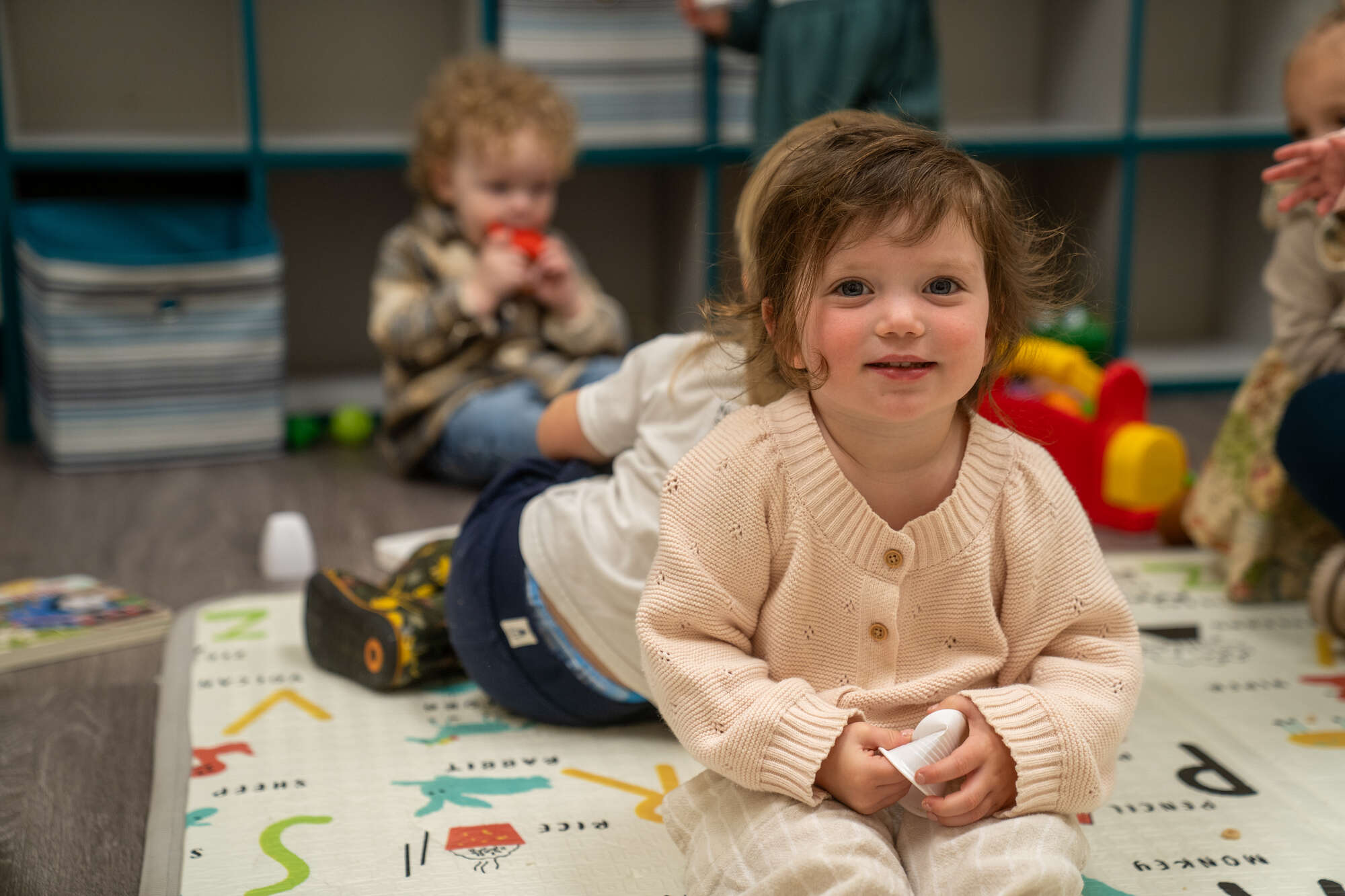 Toddler in cream sweater sitting on educational mat at church or hospital childcare program play area.