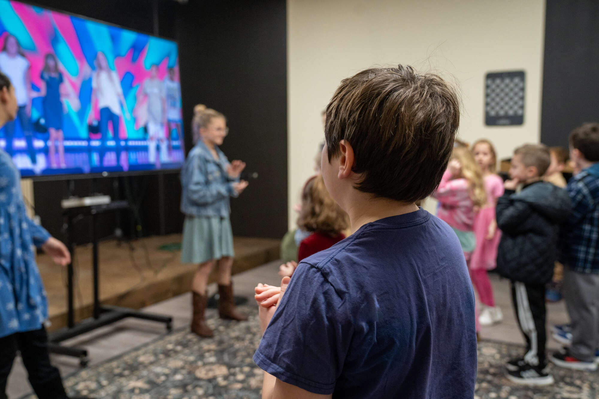 Children gathered in church, watching a religious presentation on screen with hands clasped.