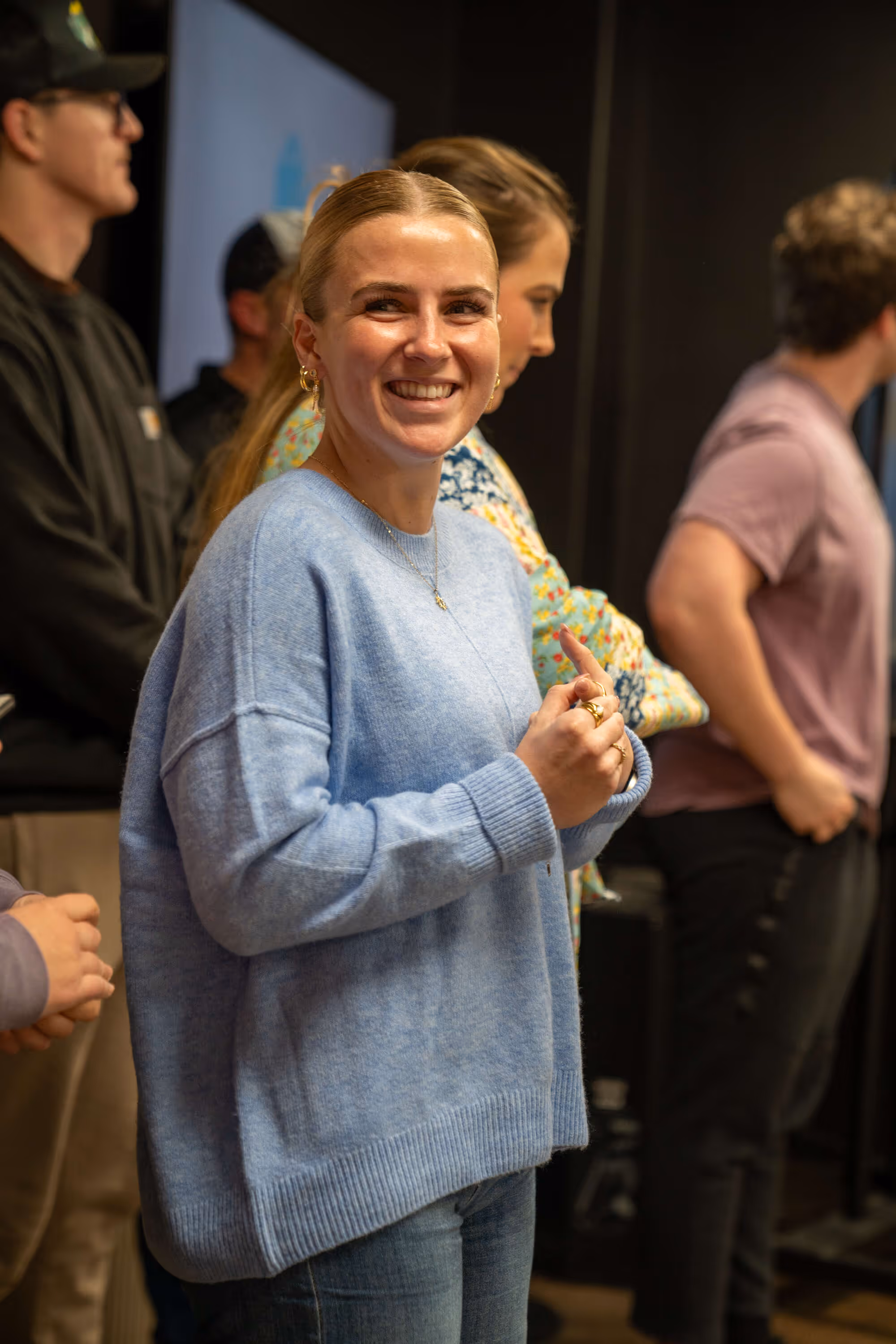Woman in blue sweater smiling with people in background at church or hospital event gathering.