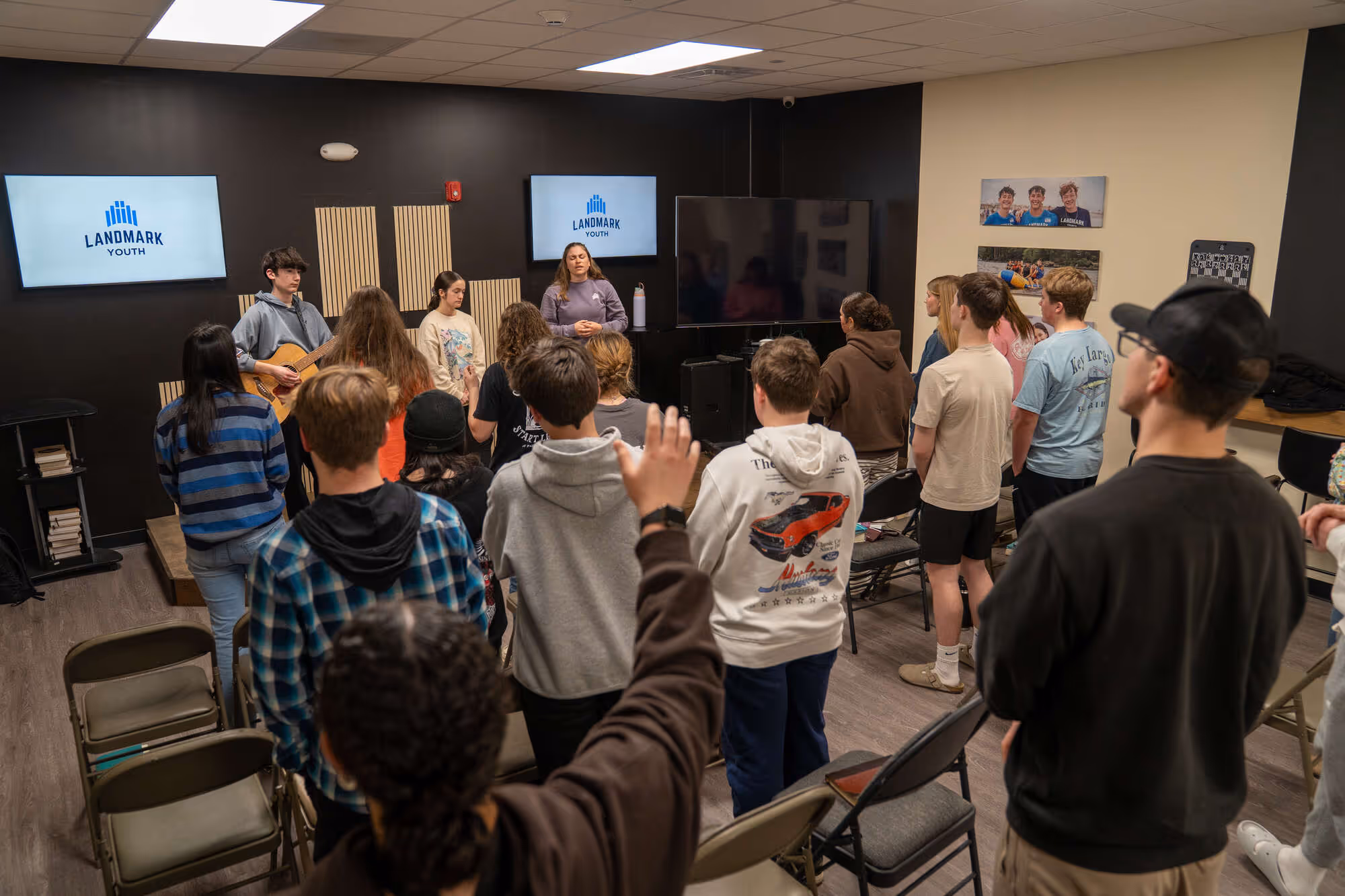Teens gathered in church room with screens displaying Landmark Youth logo, engaged in worship service.