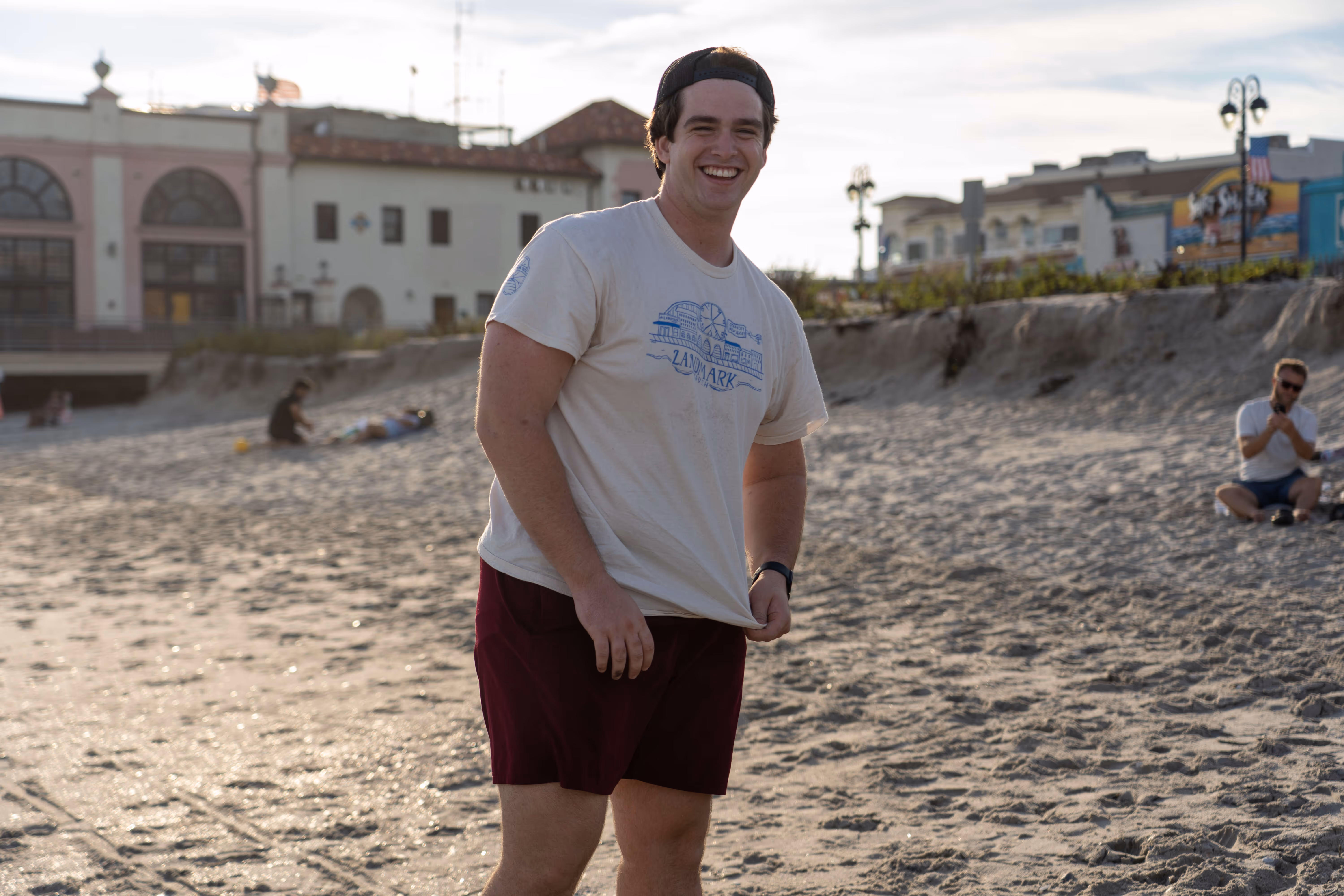 Man standing on beach wearing white t-shirt and maroon shorts smiling outdoors near buildings.