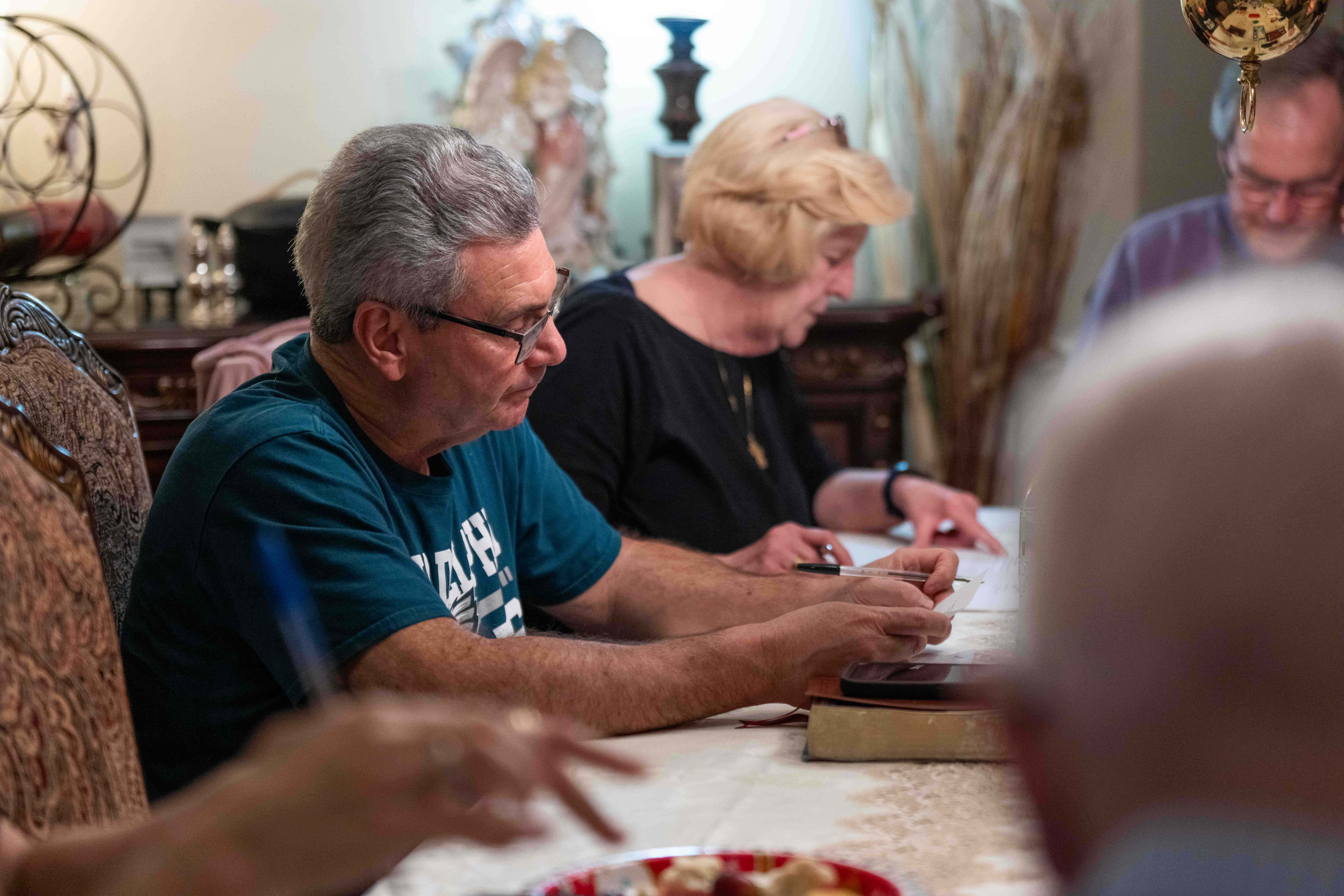 People sitting at table in church or hospital setting, engaged in activity with pen and paper.