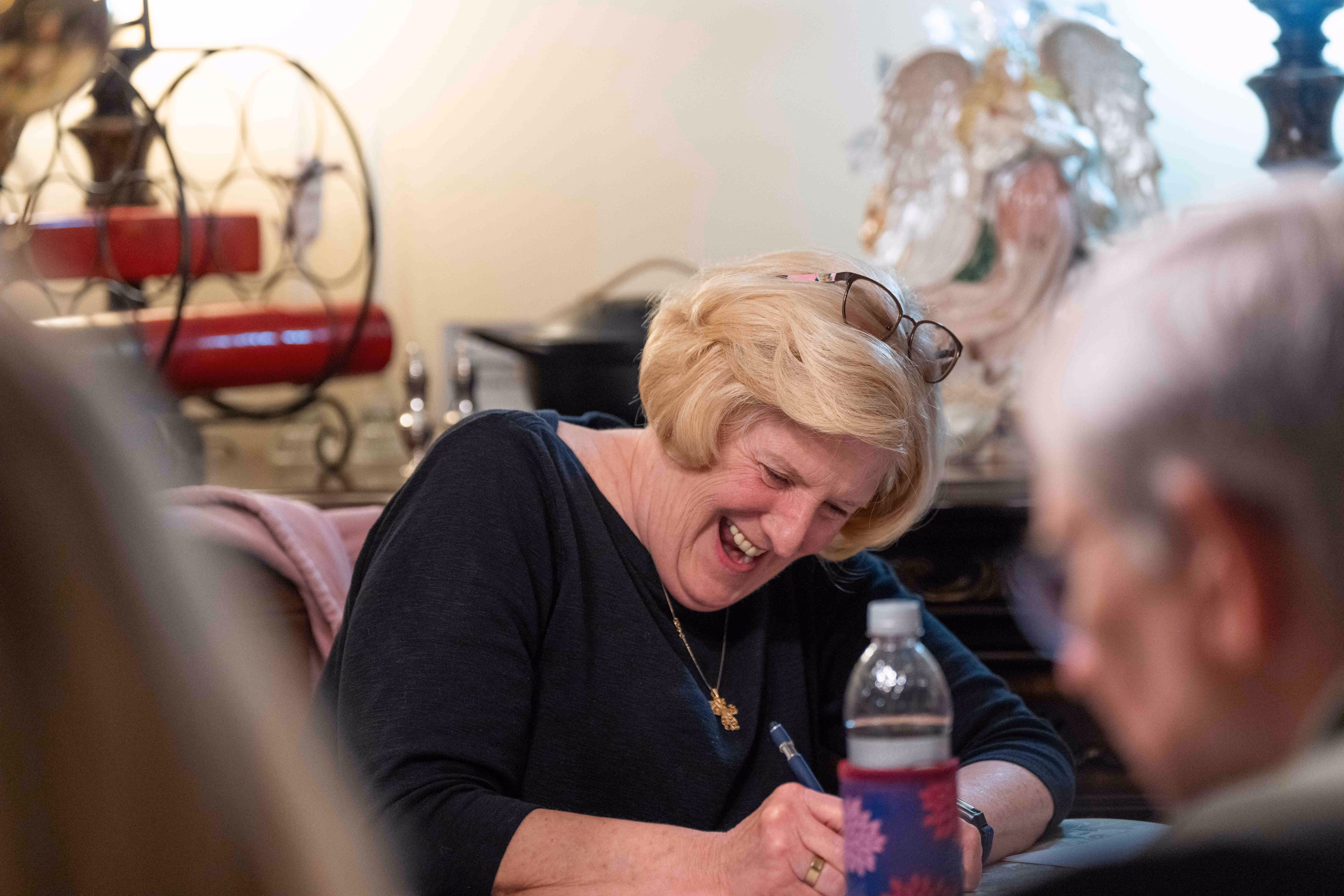 Elderly woman smiling while writing with a pen in a church or hospital setting with others.