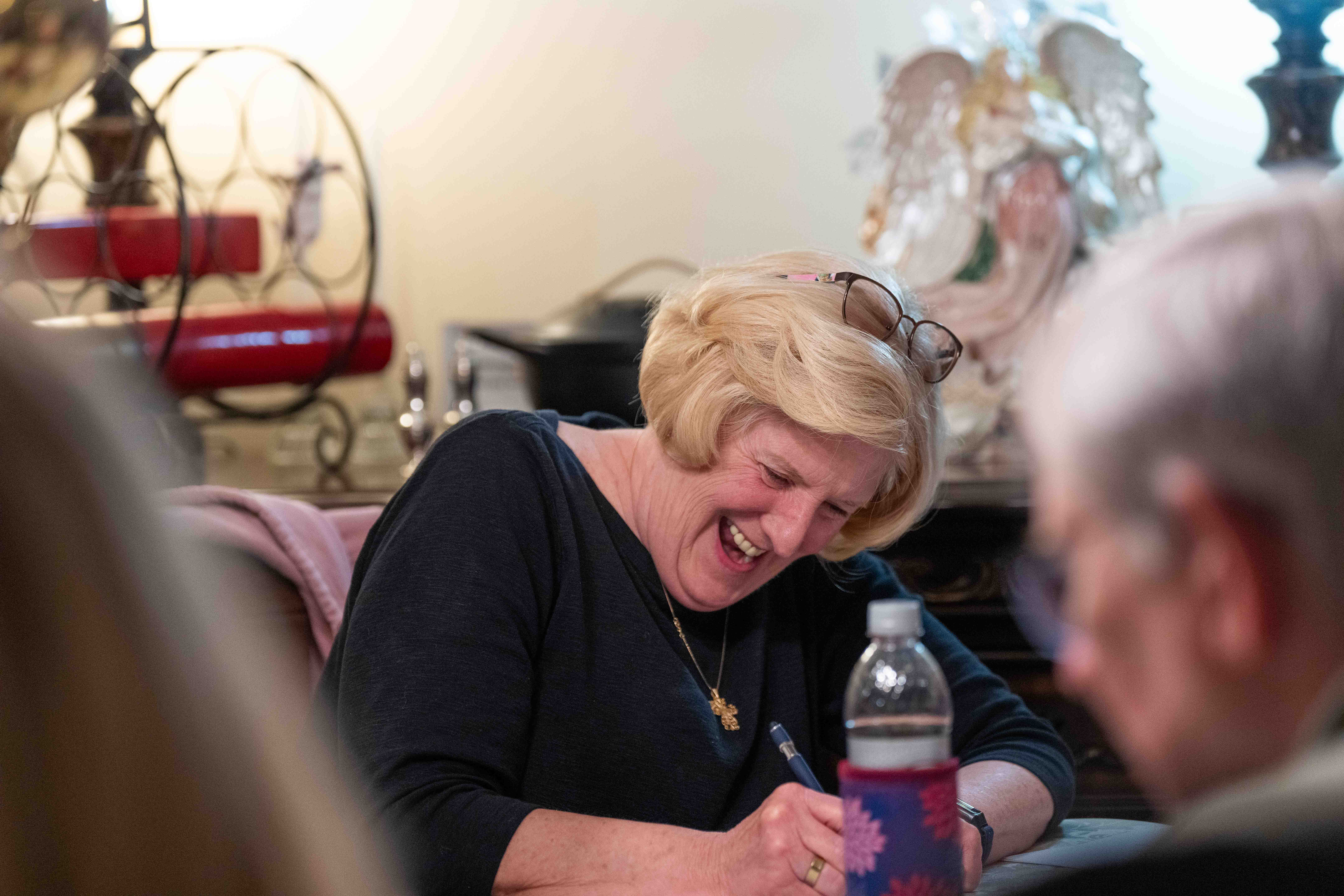 Elderly woman smiling while writing with a pen in a church or hospital setting with others.