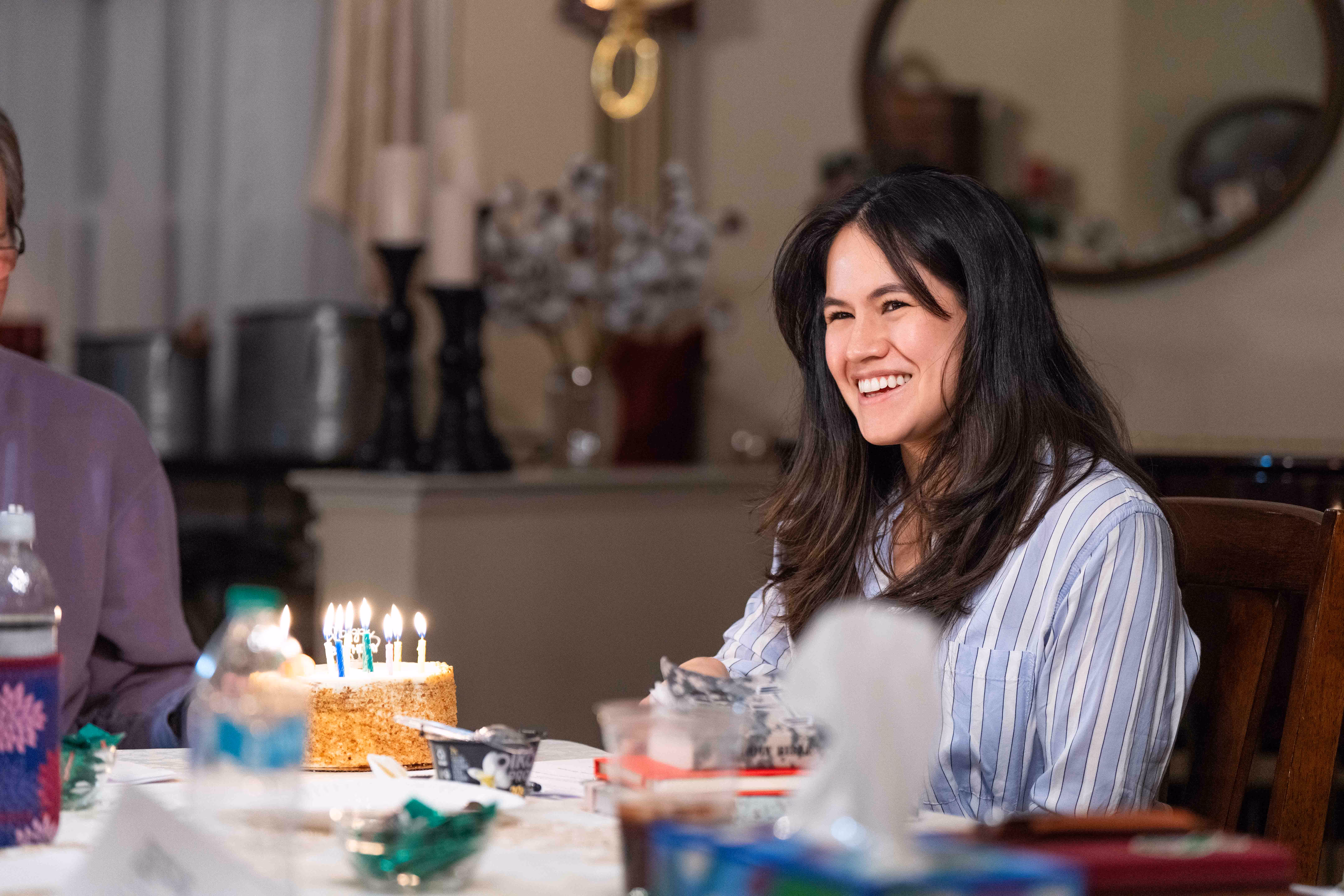 Woman sitting at table with birthday cake and candles in church or hospital setting celebration.
