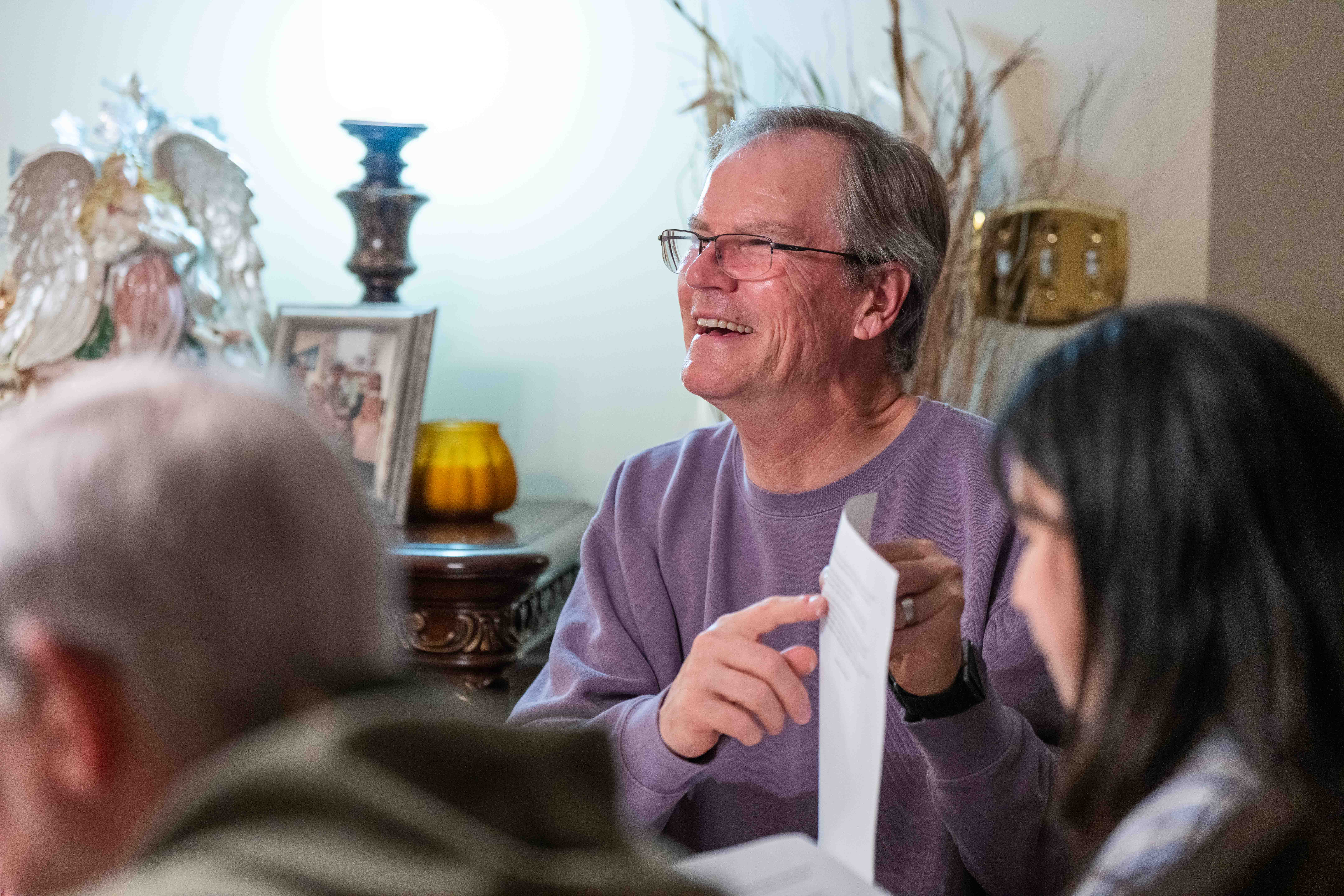 Older man in purple shirt smiling and holding paper in church or religious setting with others.