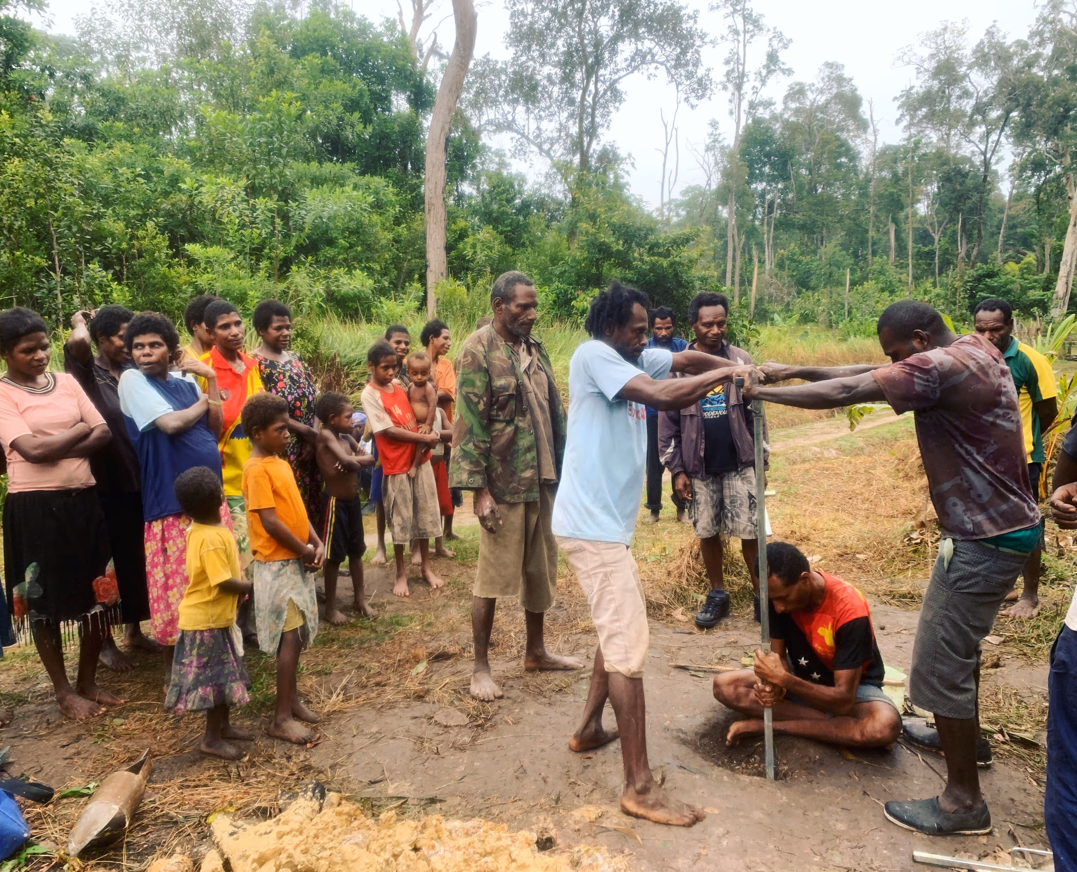 Community gathers around men demonstrating traditional craft in a rural setting near church and hospital.