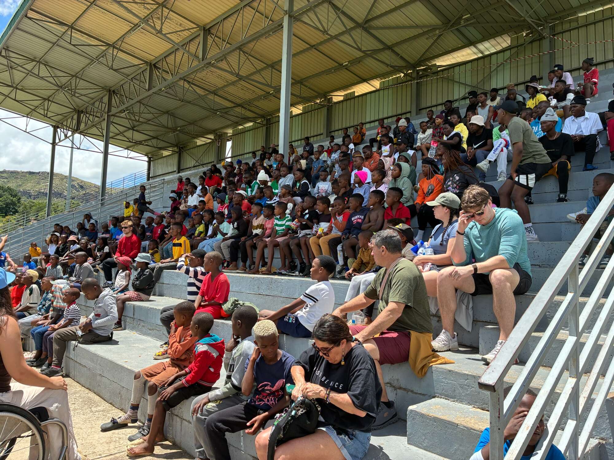 Large crowd of people sitting on bleachers at outdoor event near church or hospital gathering.
