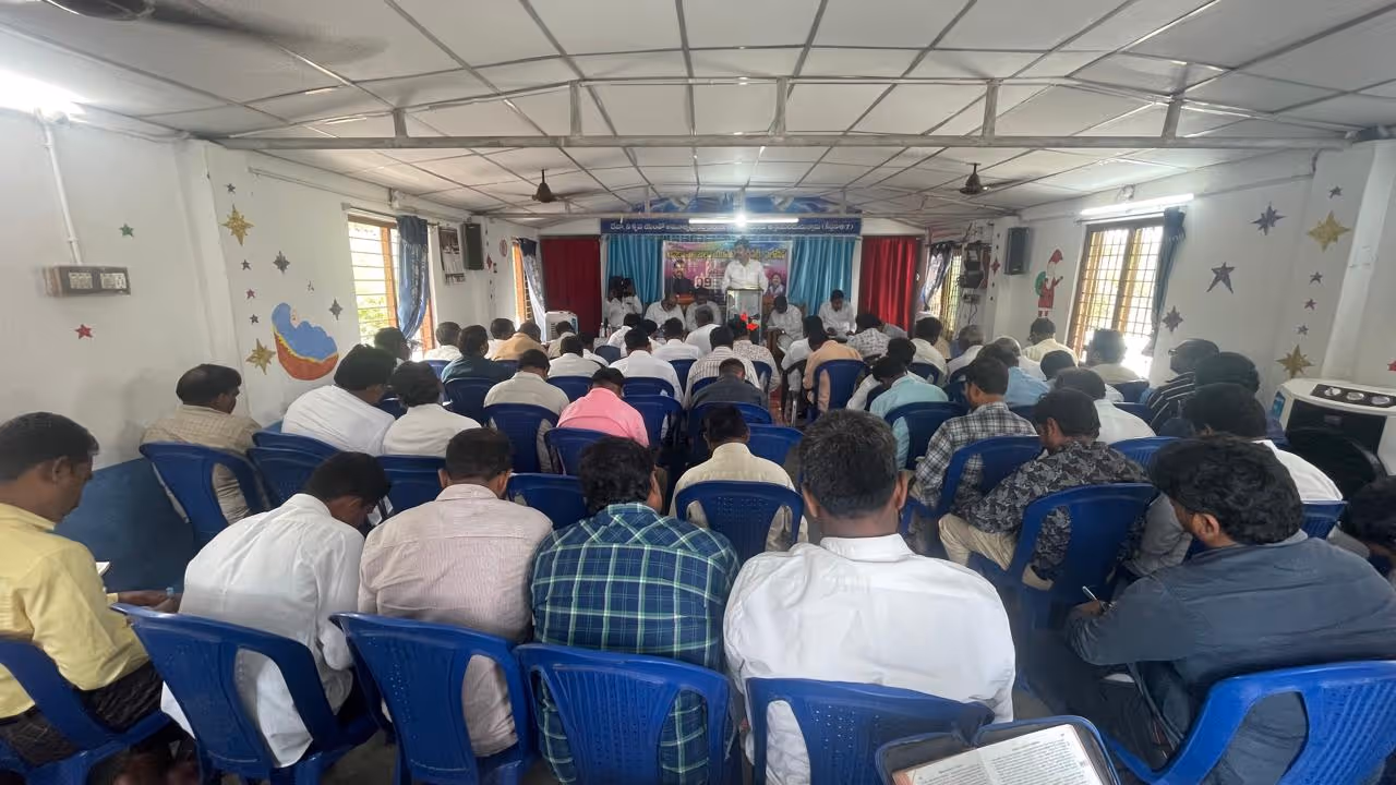 Men seated in blue chairs attending church service or religious gathering in hospital setting.