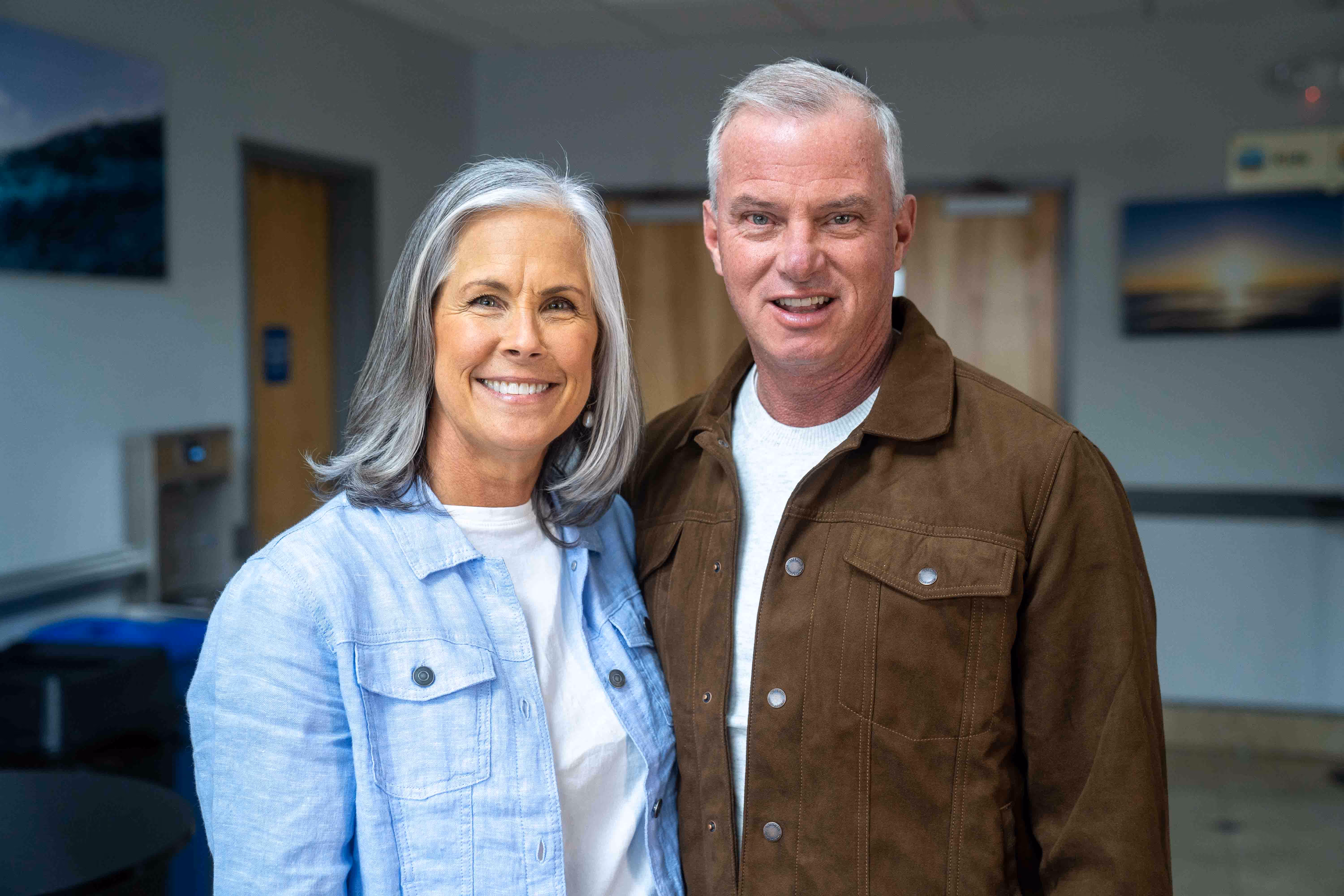 Smiling older couple indoors, woman with gray hair wearing light blue jacket, man with short gray hair wearing brown jacket.