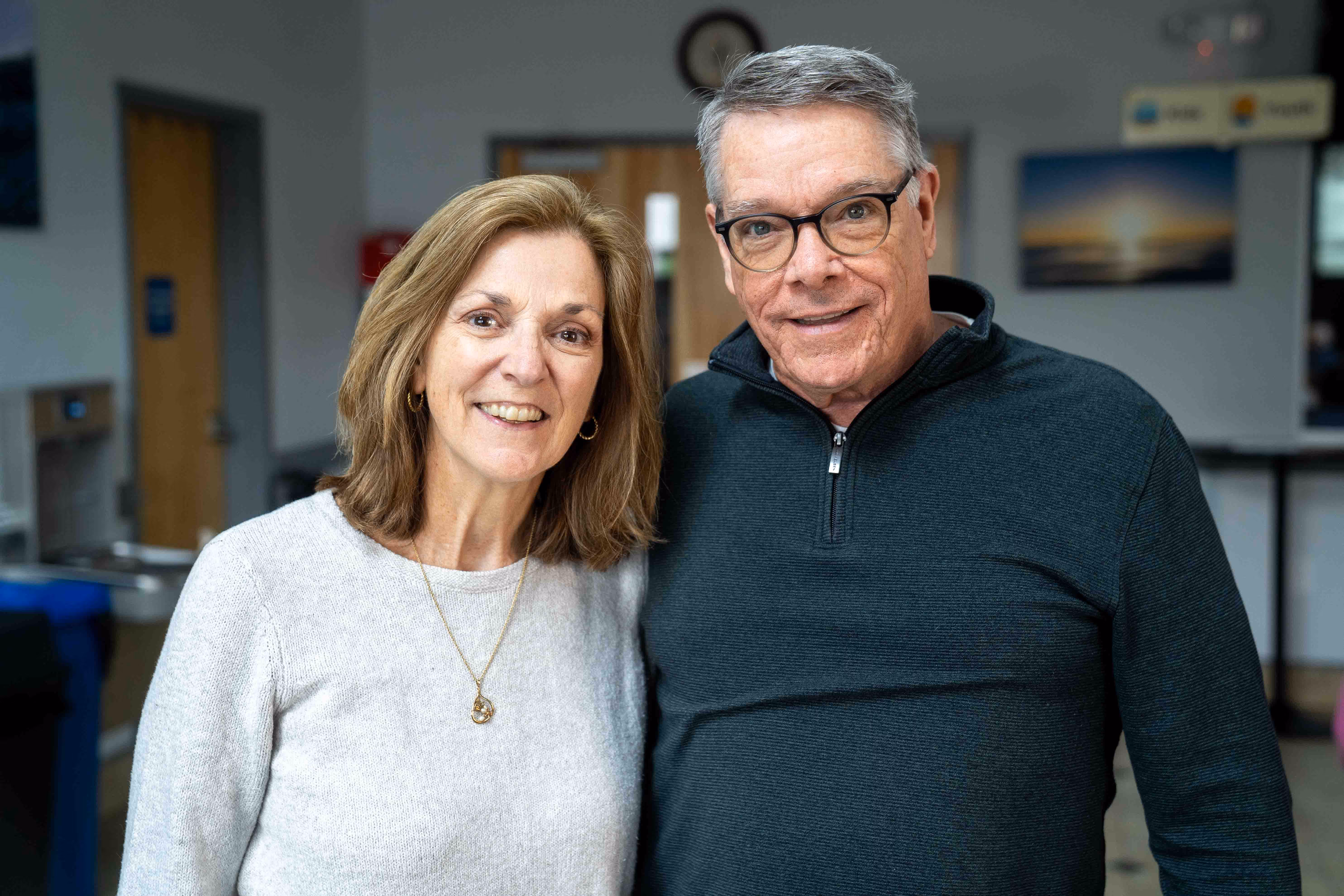 Smiling middle-aged couple posing indoors, with a woman wearing a light gray sweater and a man wearing glasses and a dark zip-up sweater.