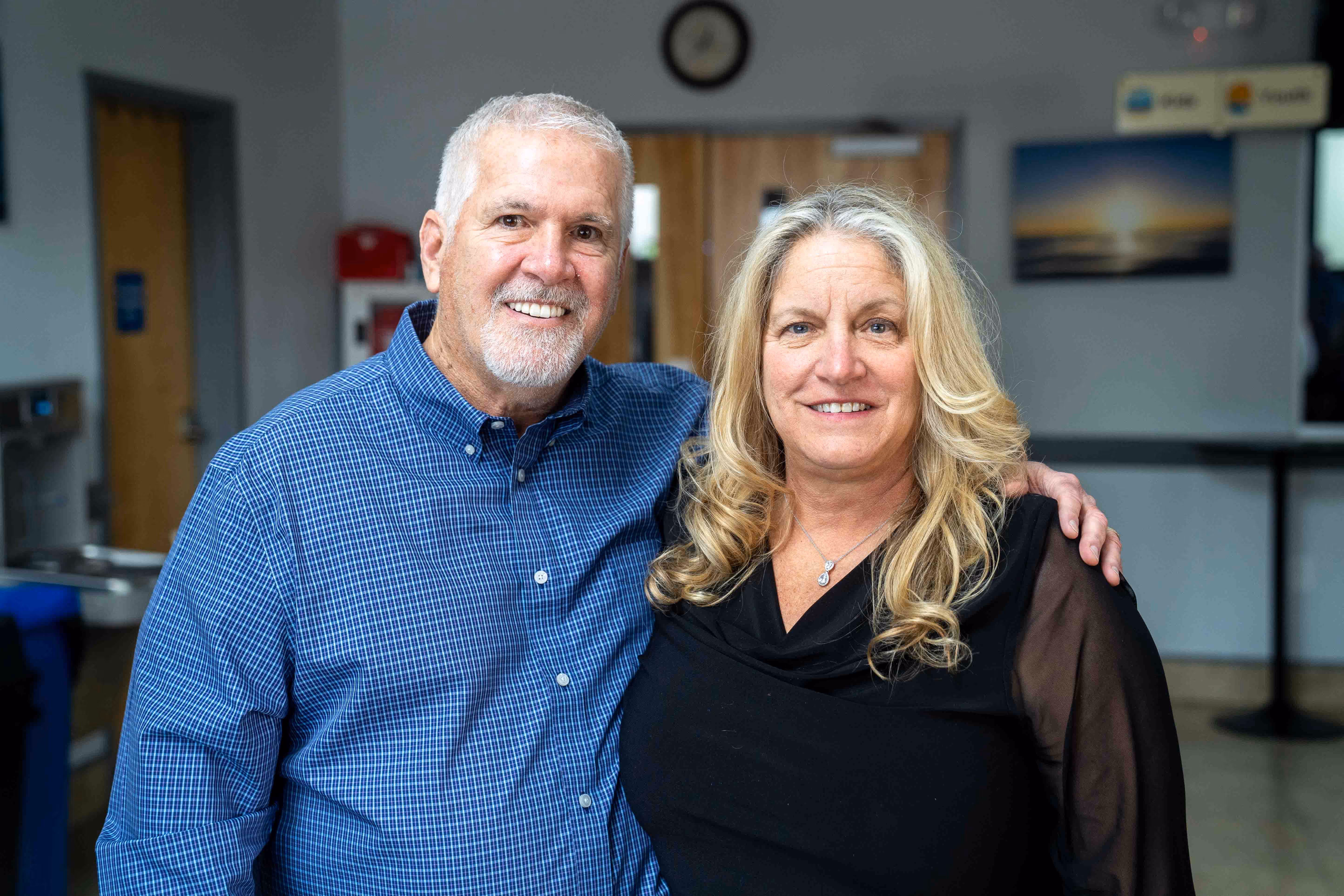 Smiling middle-aged couple with the man wearing a blue checkered shirt and the woman in a black top, standing indoors.
